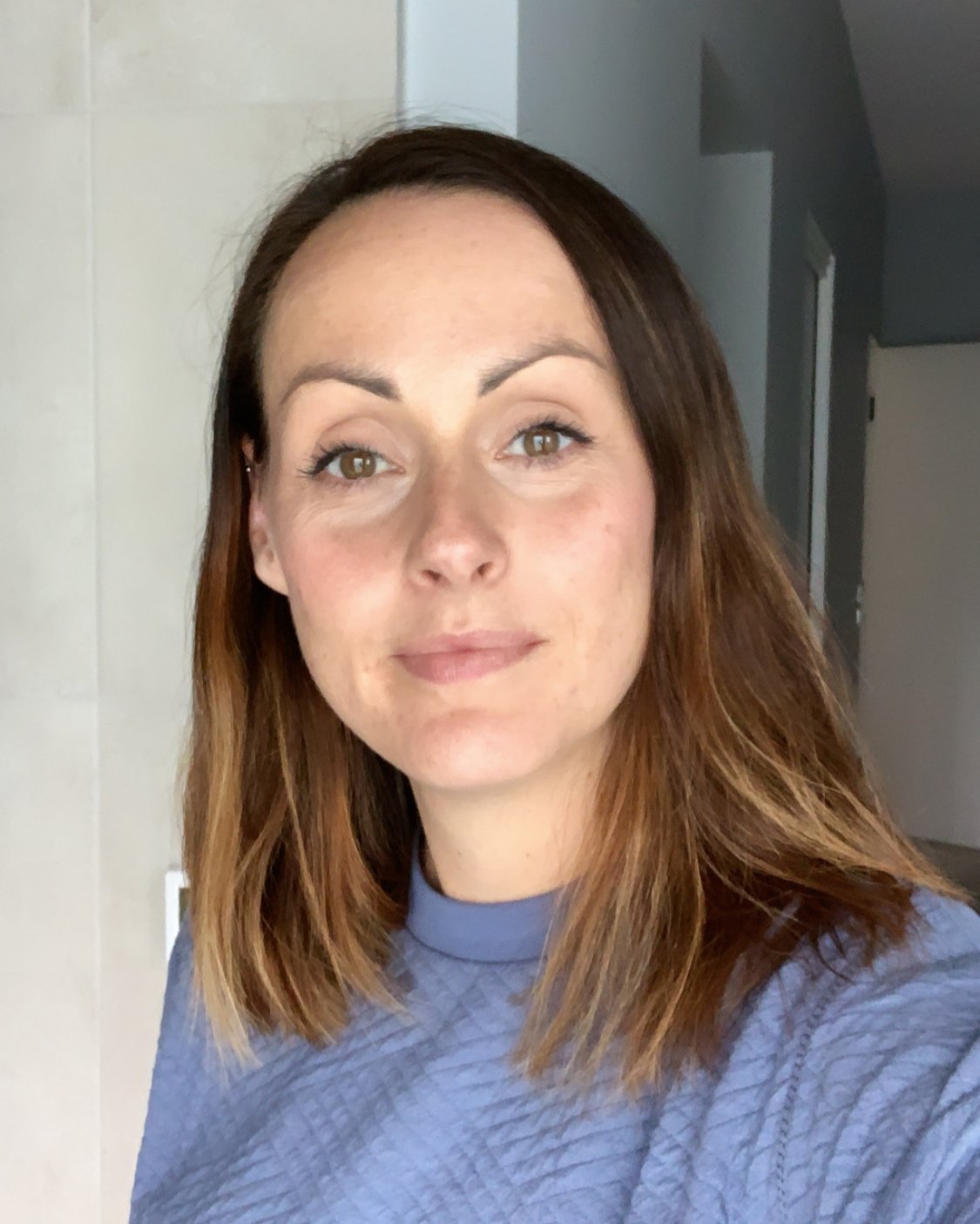 A woman with brown hair, hazel eyes, and fair skin smiling softly, wearing a light blue textured top, standing in an indoor setting.