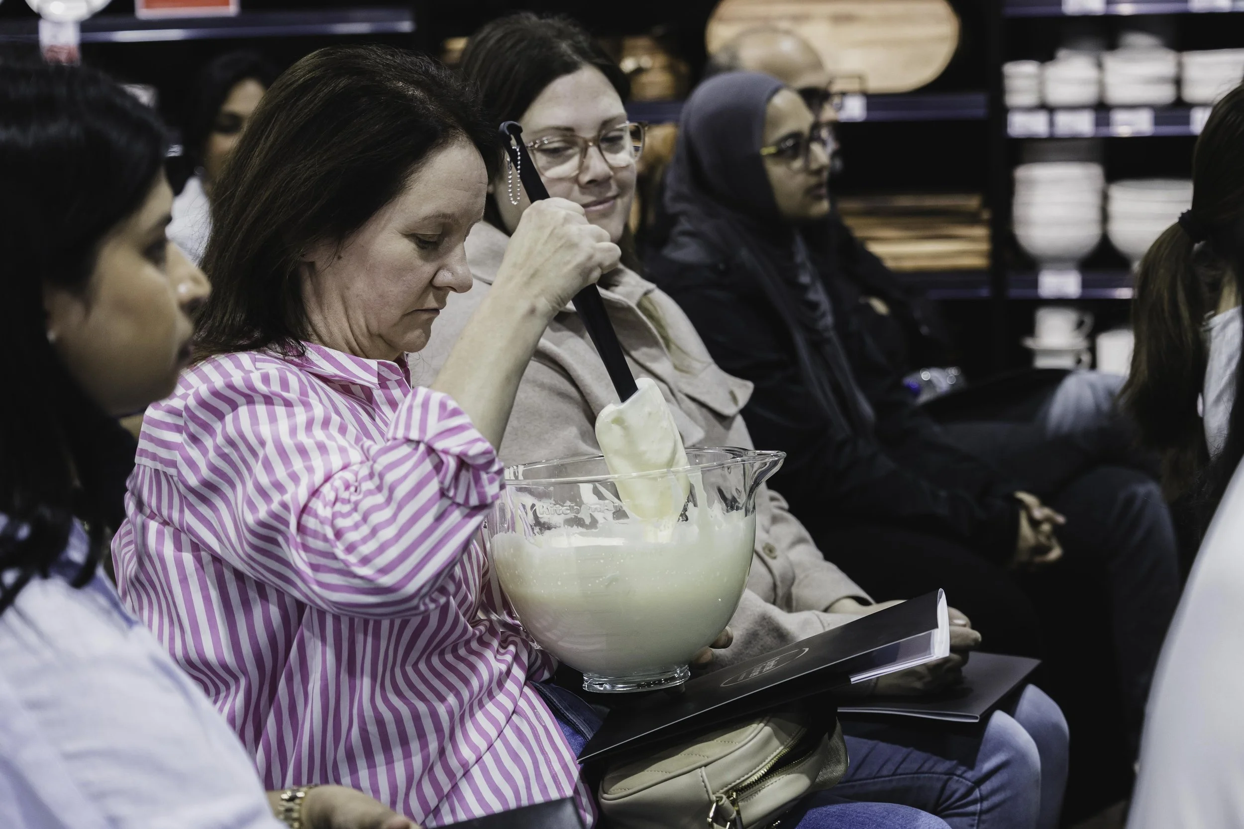 A woman in a pink and white striped shirt is sitting in a group, stirring a large glass bowl of ice cream or frozen dessert with a black spatula.