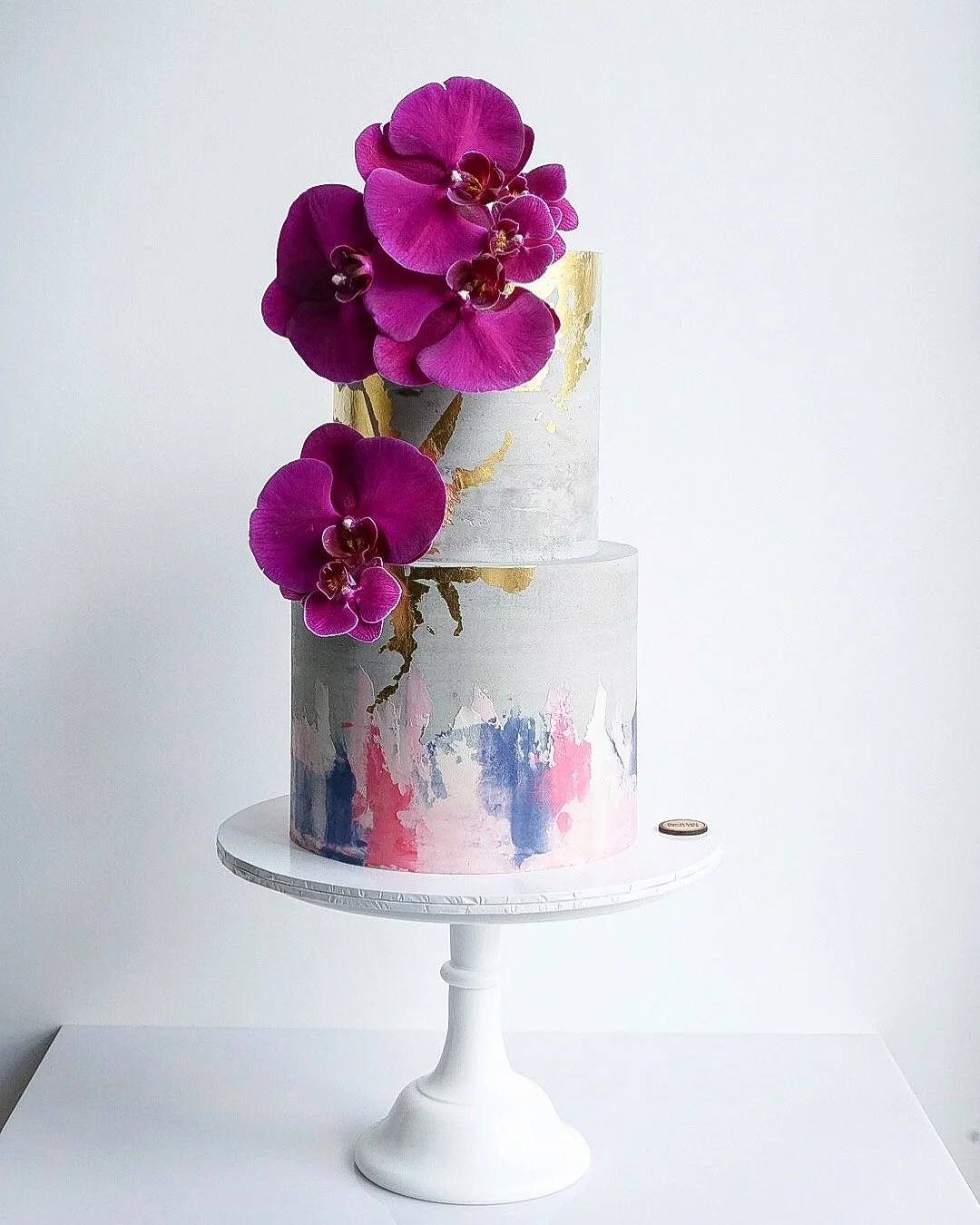 A two-tiered cake with abstract pink, blue, and gray watercolor designs, adorned with a cluster of purple orchids on top.