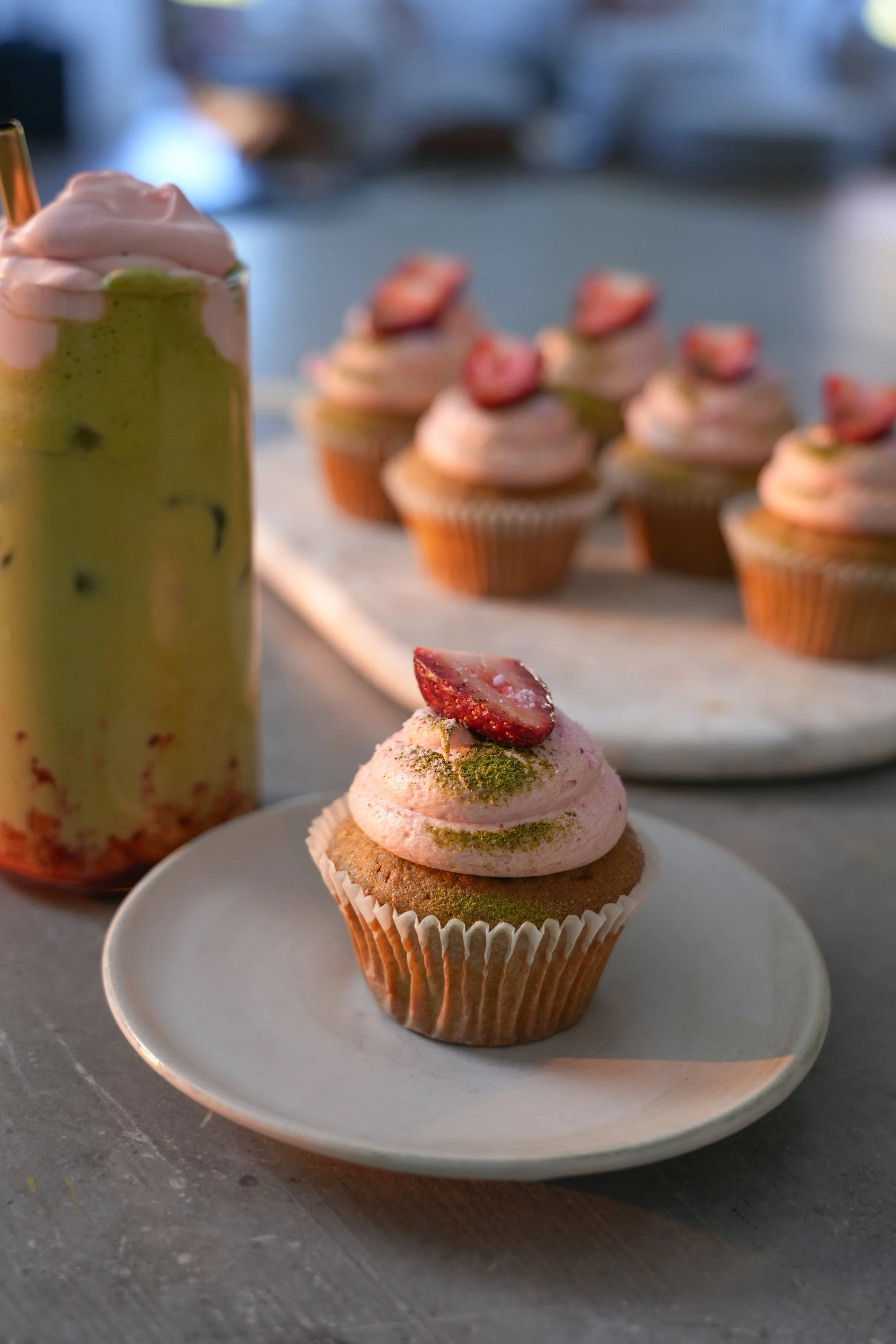 A cupcake topped with pink frosting, sliced strawberry, and green matcha powder, placed on a small white plate. In the background, there are several more cupcakes with strawberry toppings on a tray, and a green beverage with pink foam and fruit piece