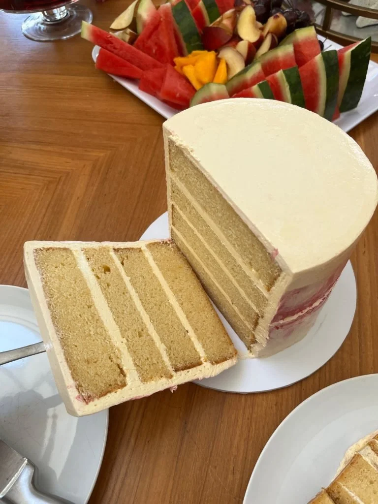 Sliced round vanilla cake with multiple layers and cream filling, placed on a white cake stand on a wooden table.