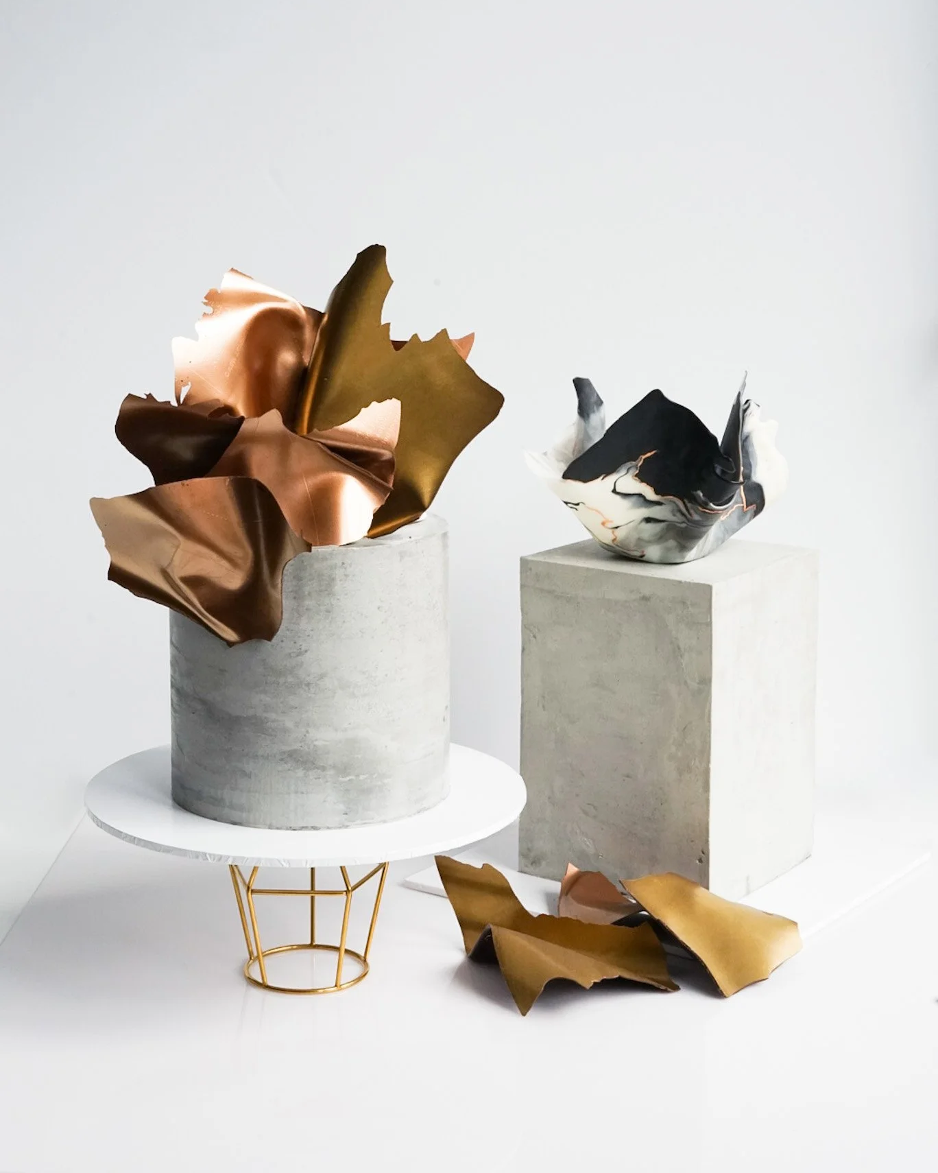 Decorative art pieces with metallic and marble-like finishes on concrete pedestals and a white surface, featuring abstract organic shapes and textures.