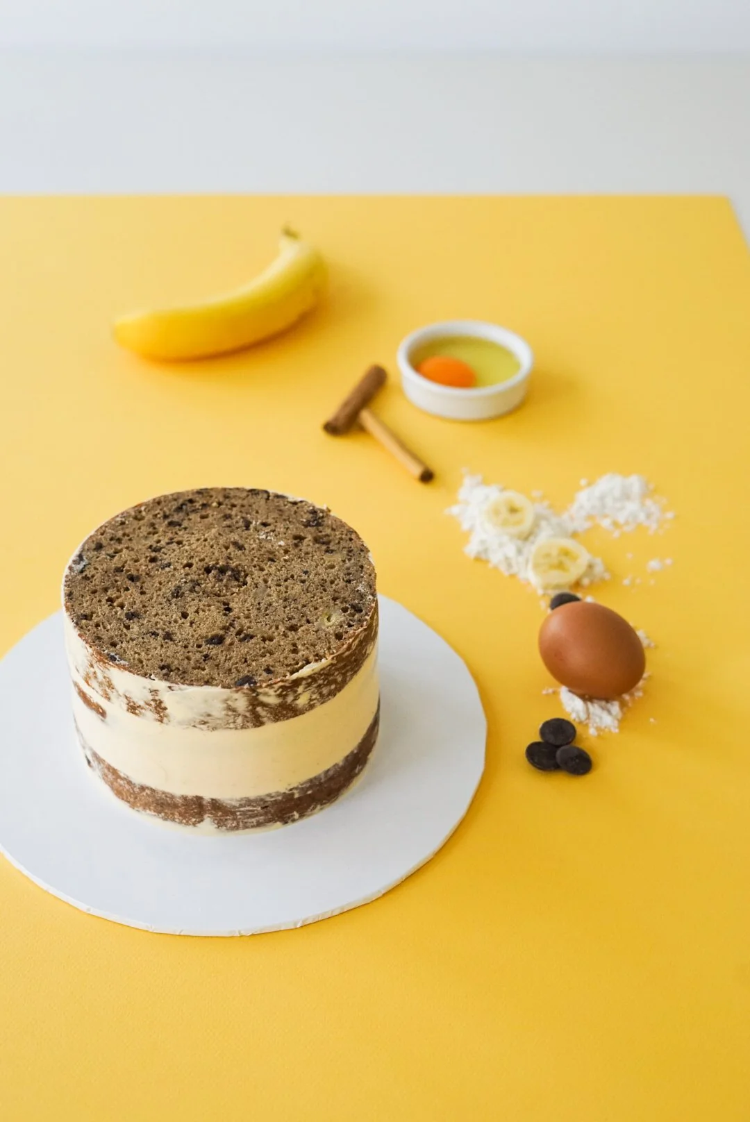 A chocolate and vanilla layered cake on a white cake board, with various baking ingredients and tools around it on a yellow background, including a banana, egg, cinnamon stick, flour, chocolate chips, and butter.