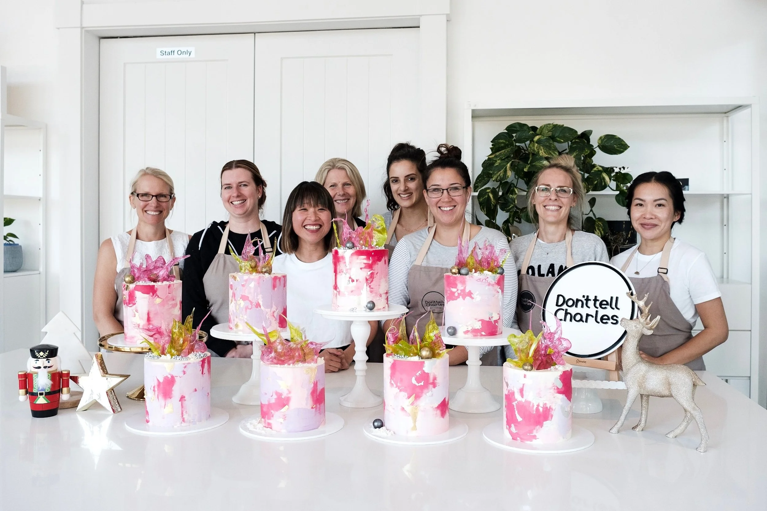Group of women celebrating a birthday with nine pink and white marbled cakes decorated with gold and pink accents, colorful foliage, and ornaments, in a bright, modern kitchen with white walls and shelving.