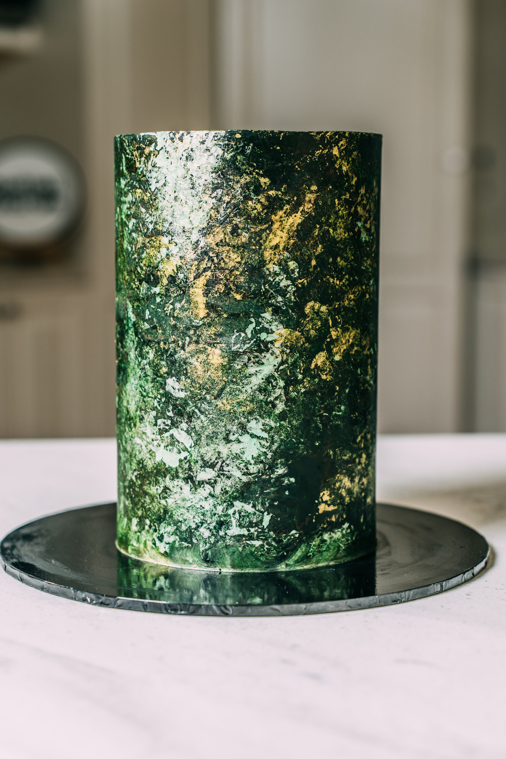 A cylindrical cake with a green, black, and gold abstract textured design on the surface, placed on a black circular base on a white table.