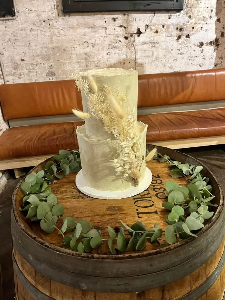 A tiered white wedding cake with dried floral decorations on top, placed on a wooden barrel with a eucalyptus garland around it in a rustic setting.