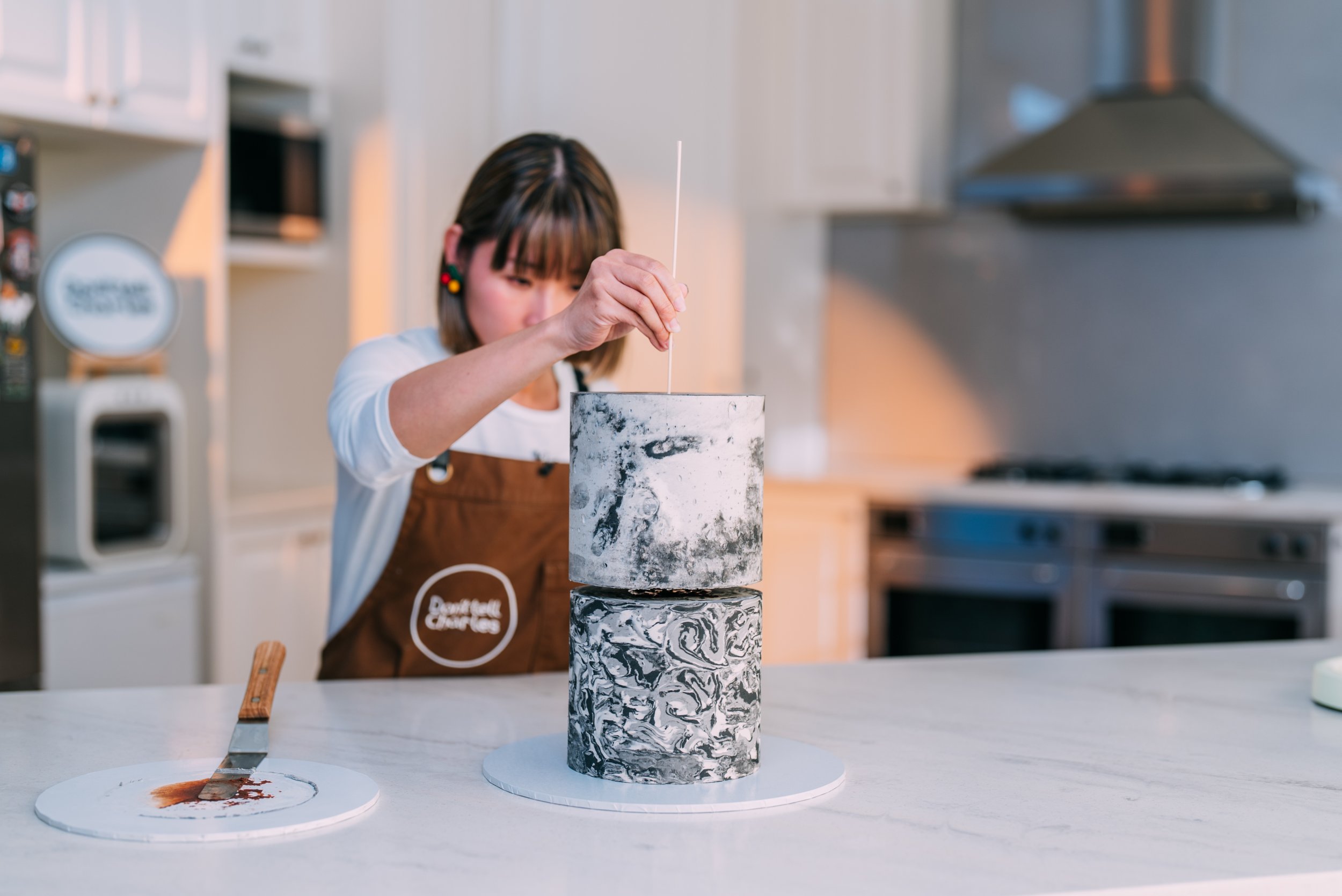 A woman wearing a brown apron with a white shirt is placing a candle wick into a tiered, marble-patterned cake on a kitchen countertop.
