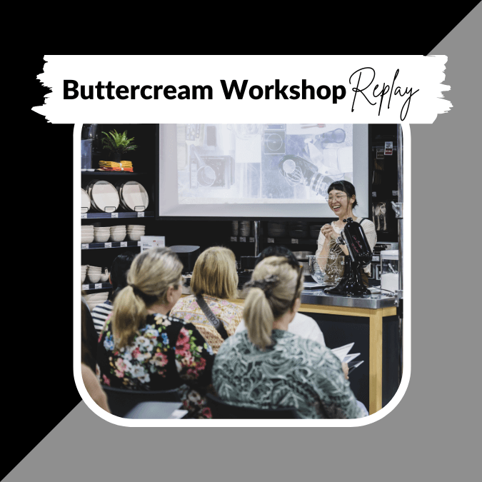 A woman in a black apron demonstrates how to make buttercream icing at a workshop, with an audience of six women watching intently in a kitchen setting with shelves of dishes and a large screen in the background.