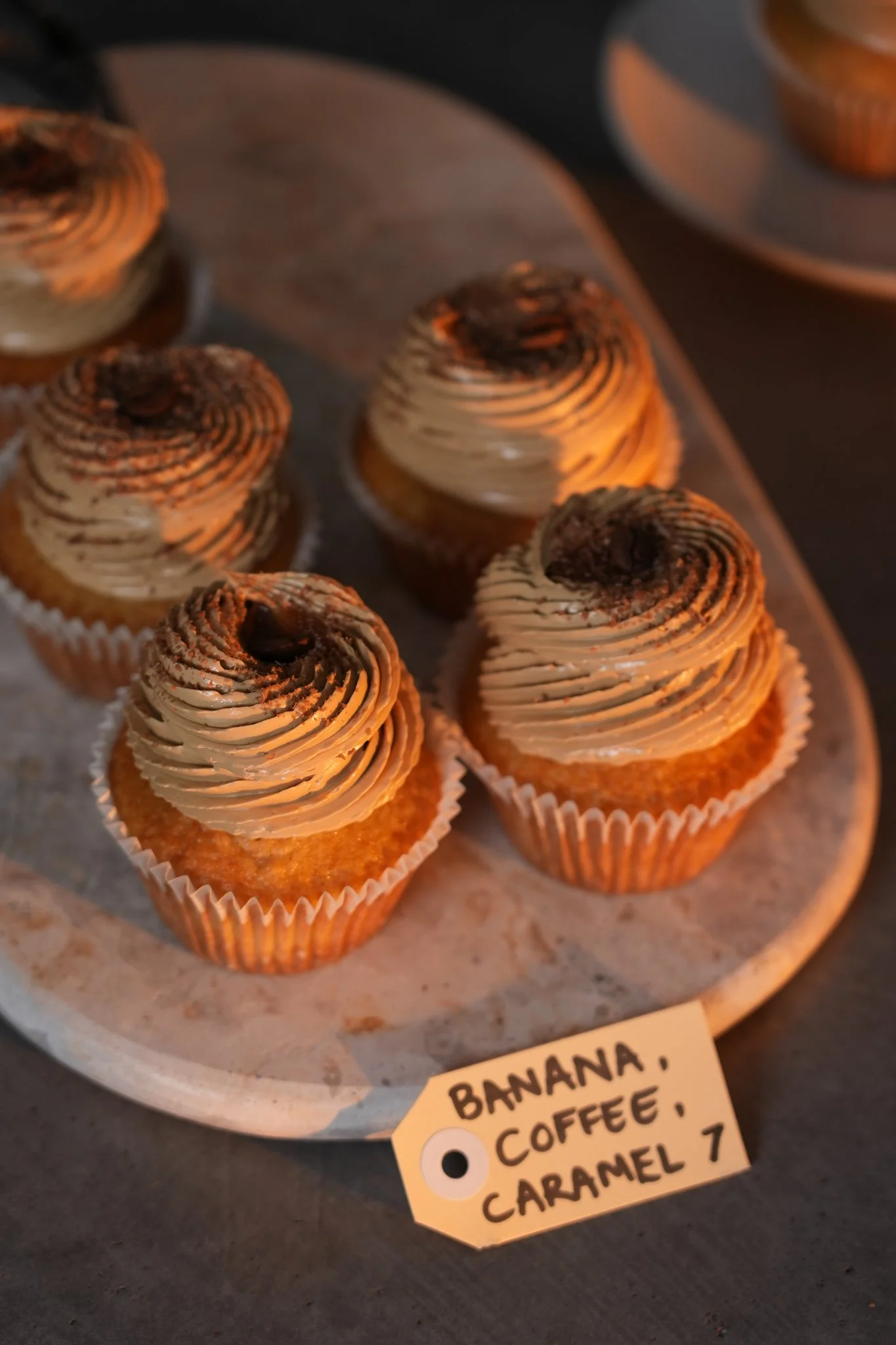 A tray of banana cupcakes with coffee and caramel flavors, topped with swirls of frosting and a sprinkle of chocolate, labeled "BANANA, COFFEE, CARAMEL 7."