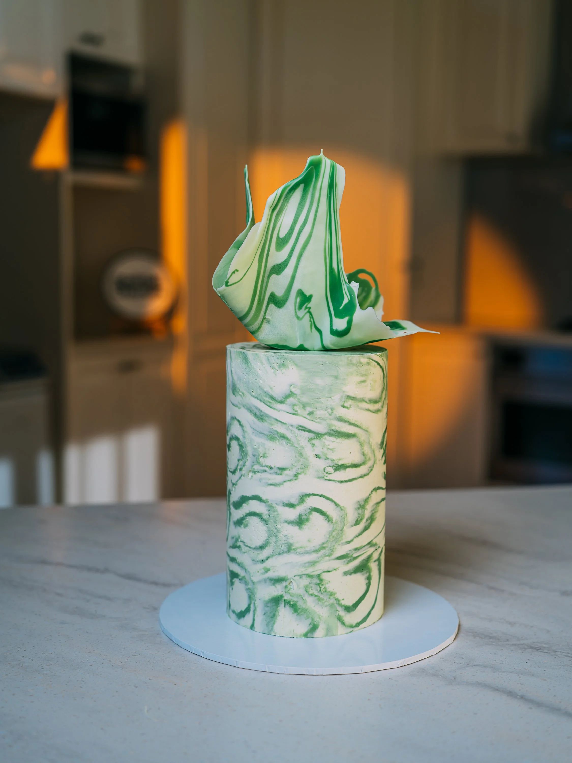 A cake with white and green marbled fondant decorations, topped with a green artful sugar sculpture, on a white circular cake board placed on a kitchen countertop.