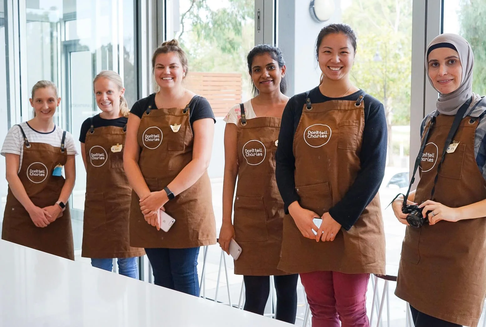 Group of six women standing indoors, wearing brown aprons with a logo, smiling, some holding phones and a camera, in front of large windows with trees outside.