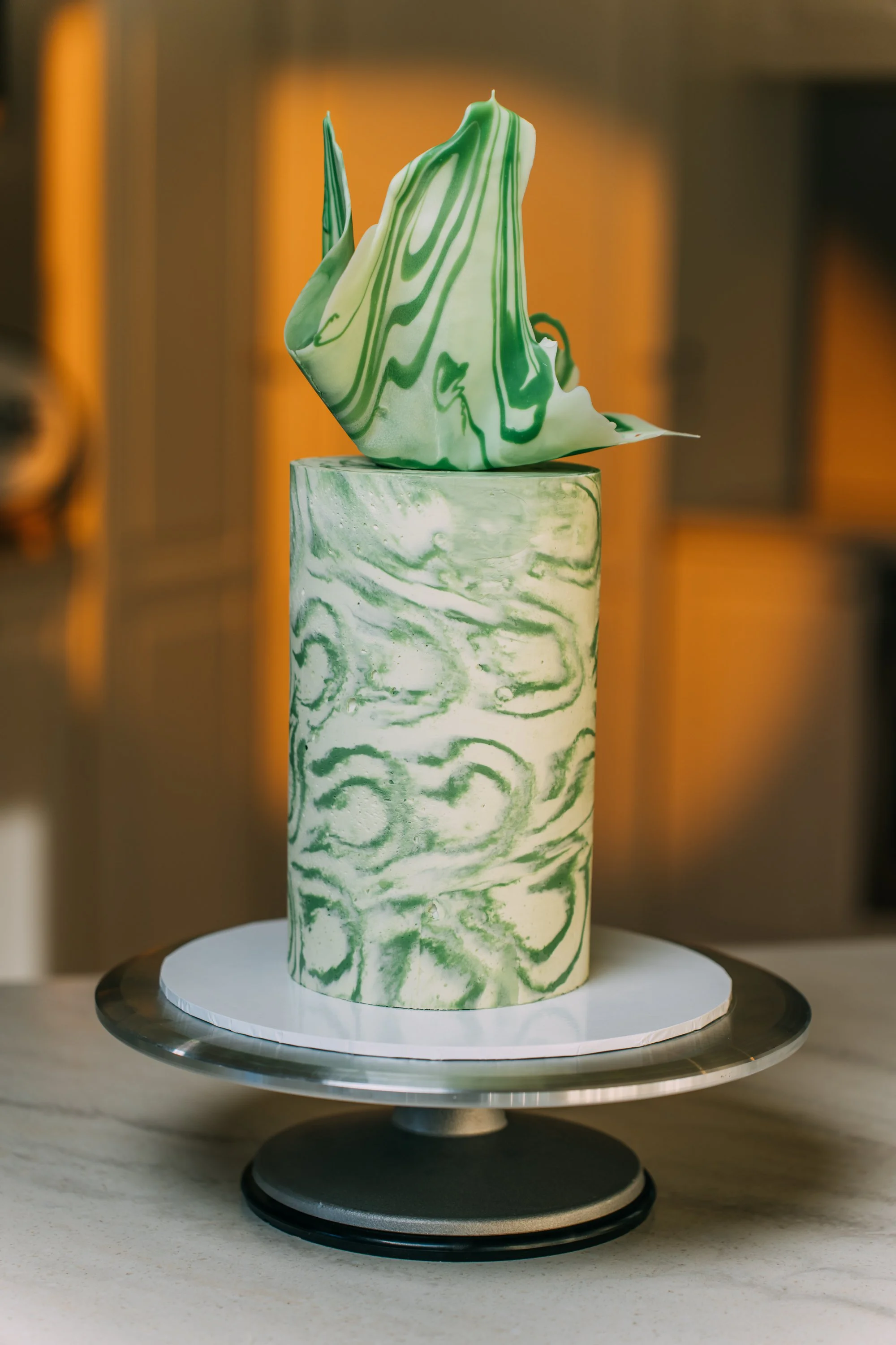 A tall cake with green and white marbled patterns, topped with a green and white marbled abstract sculpture, on a white and silver cake stand.