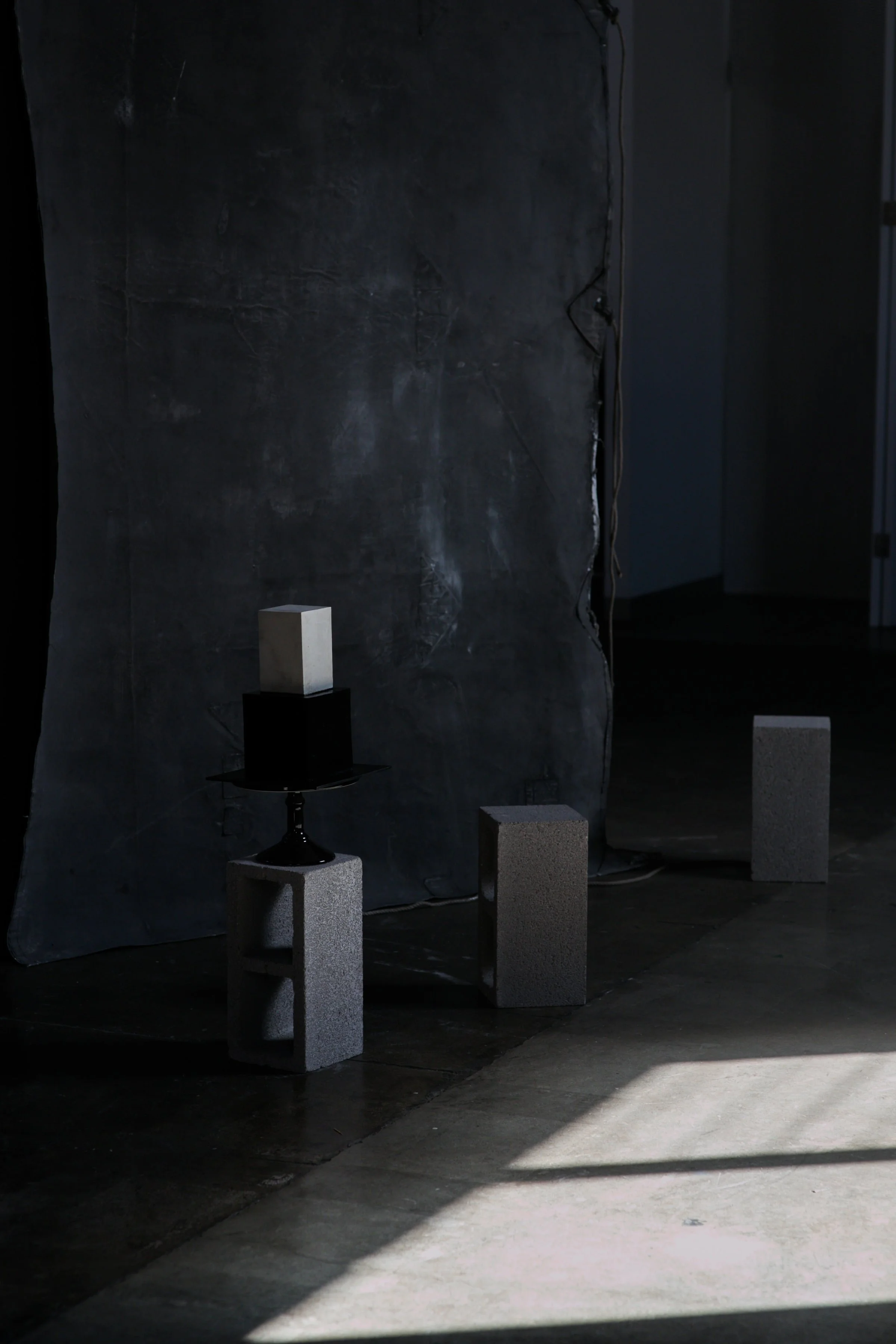 Art installation with three cinder blocks, a small black table, and a rectangular sculpture against a textured dark wall, with sunlight casting shadows on the floor.