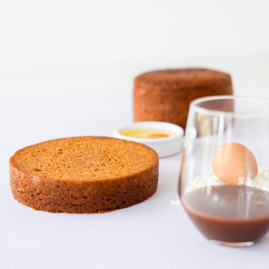 A slice of chocolate cake, a whole chocolate cake in the background, a glass of chocolate milk, and a small bowl of caramel sauce on a white surface.
