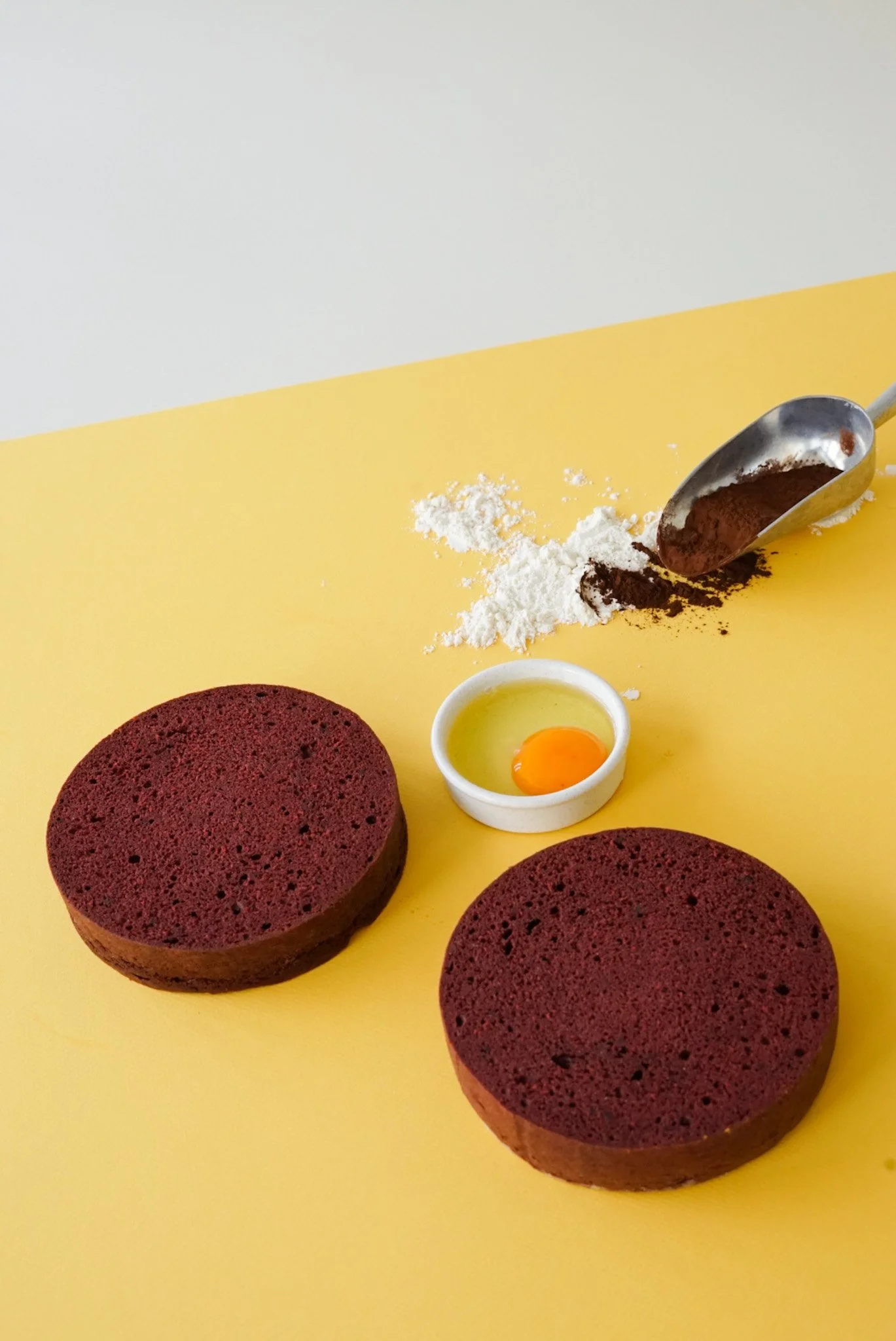 Two round pans of red velvet cake on a yellow surface, with a small dish of egg yolk, a scoop of cocoa powder, and flour nearby, suggesting cake baking preparations.