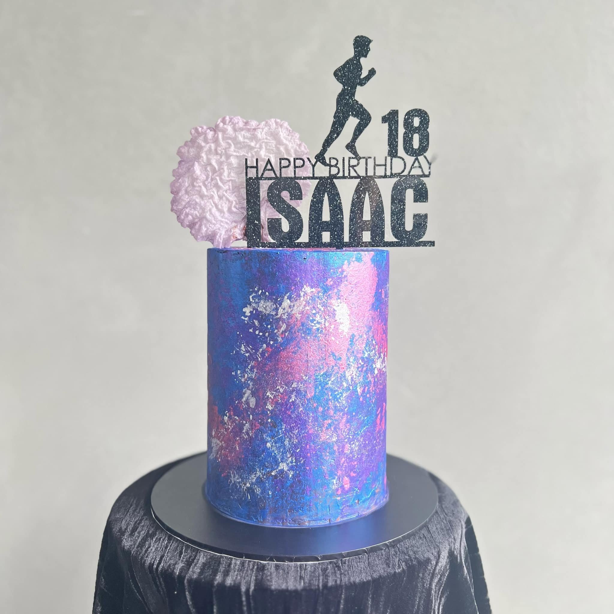 Holographic birthday cake with a black topper that reads 'Happy Birthday Isaac', '18', a silhouette of a running figure, and 'Love.' A pink cotton candy decoration is on the side of the topper. The cake is on a black textured stand.