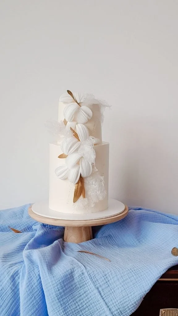 A tall white cake decorated with white floral designs and brown leaves, placed on a wooden cake stand with a light blue cloth underneath.