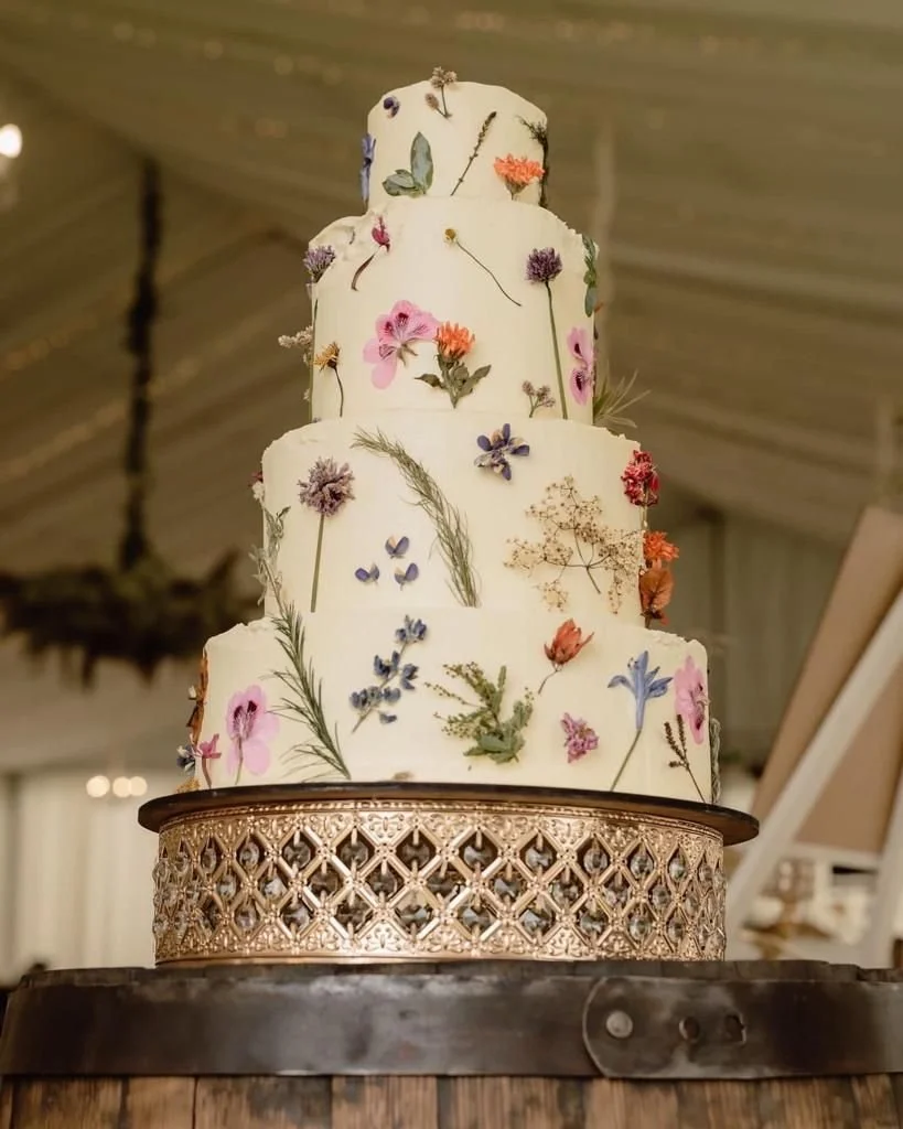 A four-tier wedding cake decorated with edible and real dried flowers and herbs, placed on a decorative gold stand on a wooden surface.