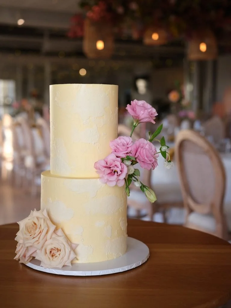 Two-tiered pale yellow wedding cake decorated with pink and white flowers, placed on a wooden table in a decorated venue.