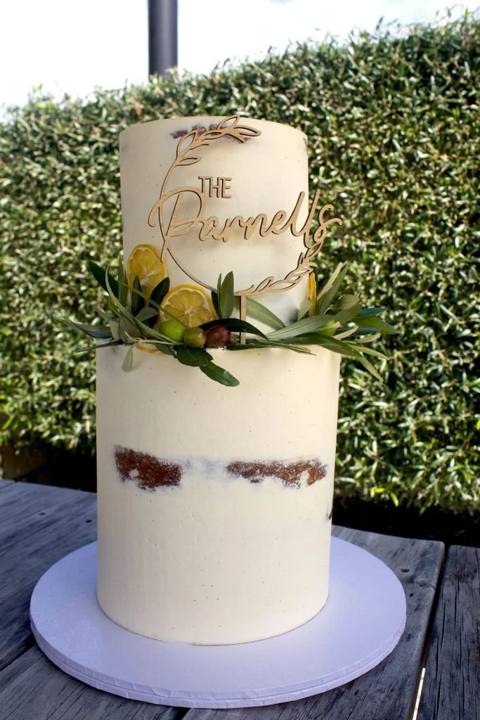 A tall wedding cake with white frosting, decorated with lemon slices, green olives, and greenery. A floral cake topper reads 'The Barnels'.