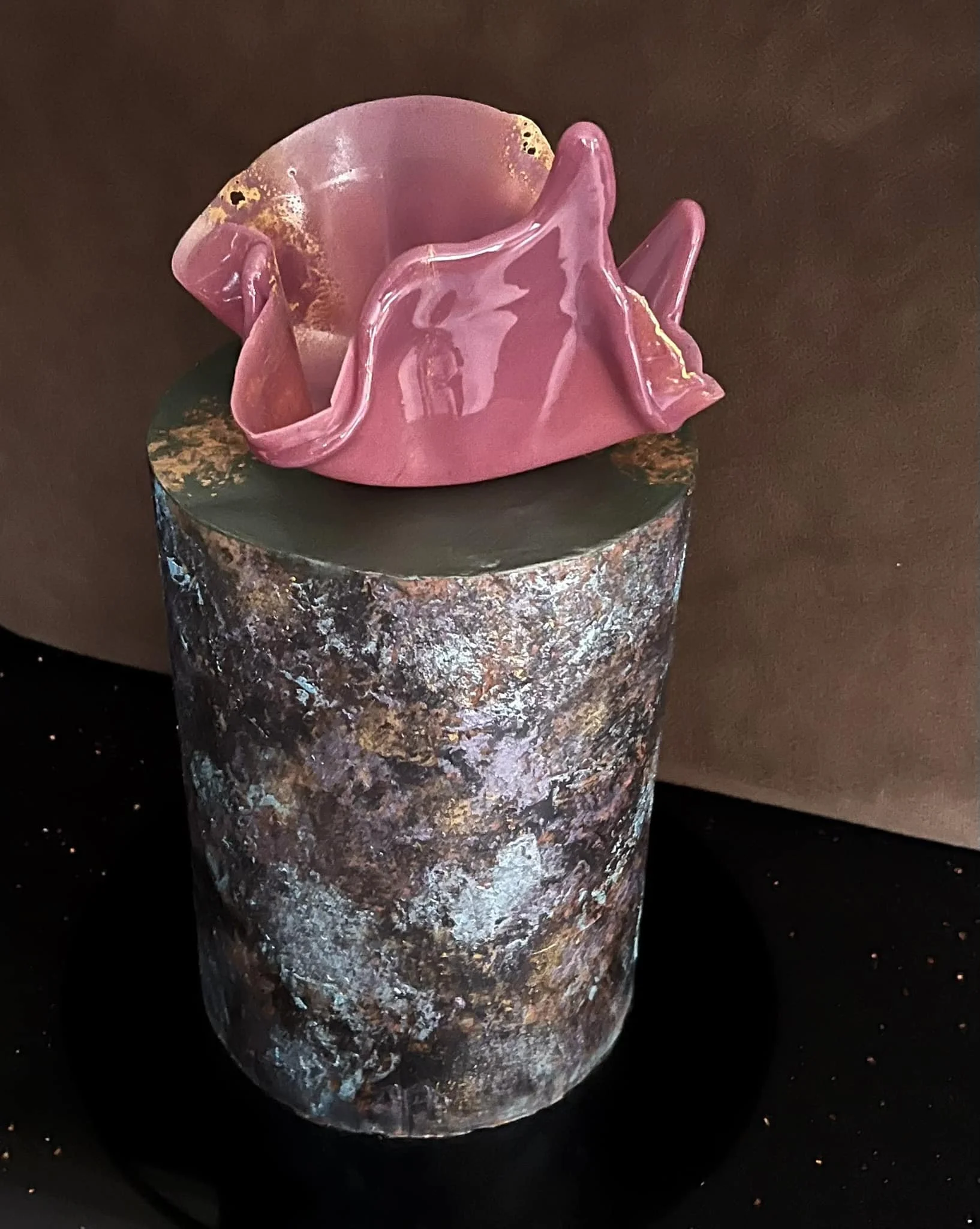 A decorative vase with a marbled, textured surface in shades of brown, gray, and tan, topped with an abstract pink glass sculpture that has a smooth, flowing form with glossy finish, set against a dark background.