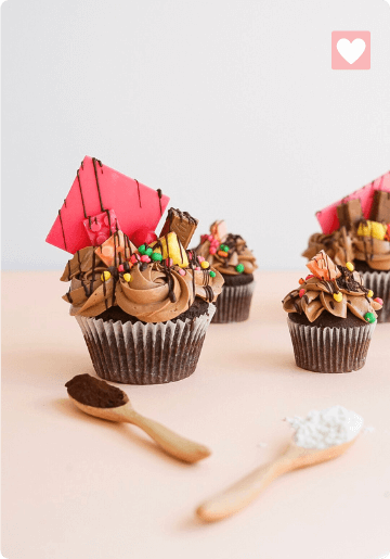 Chocolate cupcakes decorated with colorful candies, wafers, and pink chocolate shards, with two spoons of sprinkles on the side.