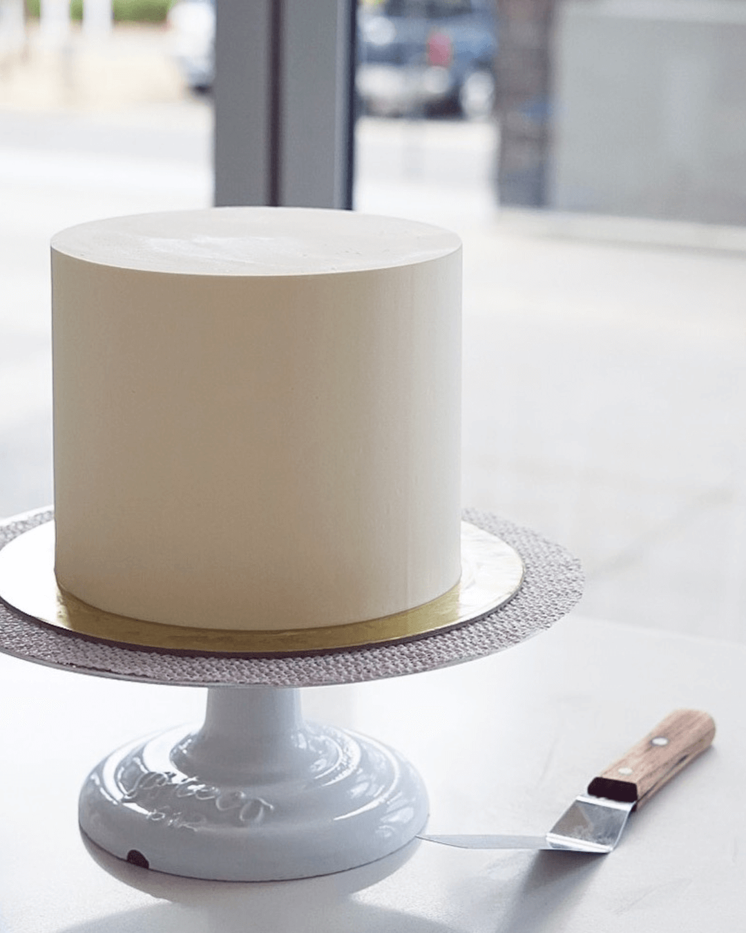 A white, cylindrical cake on a cake stand, with a cake spatula beside it, inside a bakery or cafe.