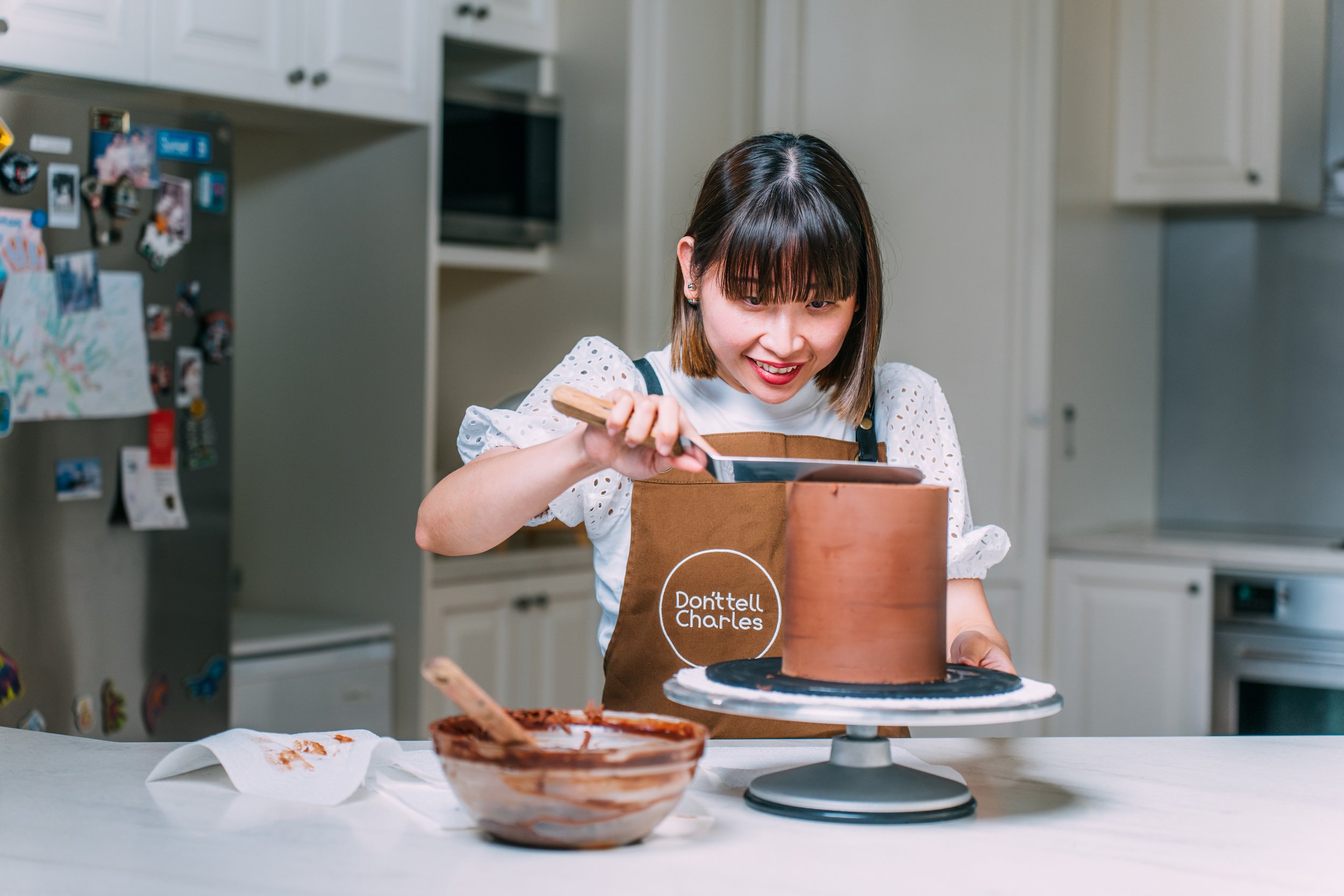 A woman in a kitchen decorating a chocolate cake while smiling. She is wearing an apron with the text 'Don't tell Charles' and using a small offset spatula. There is a bowl of chocolate frosting and a paper towel with some frosting on the counter.