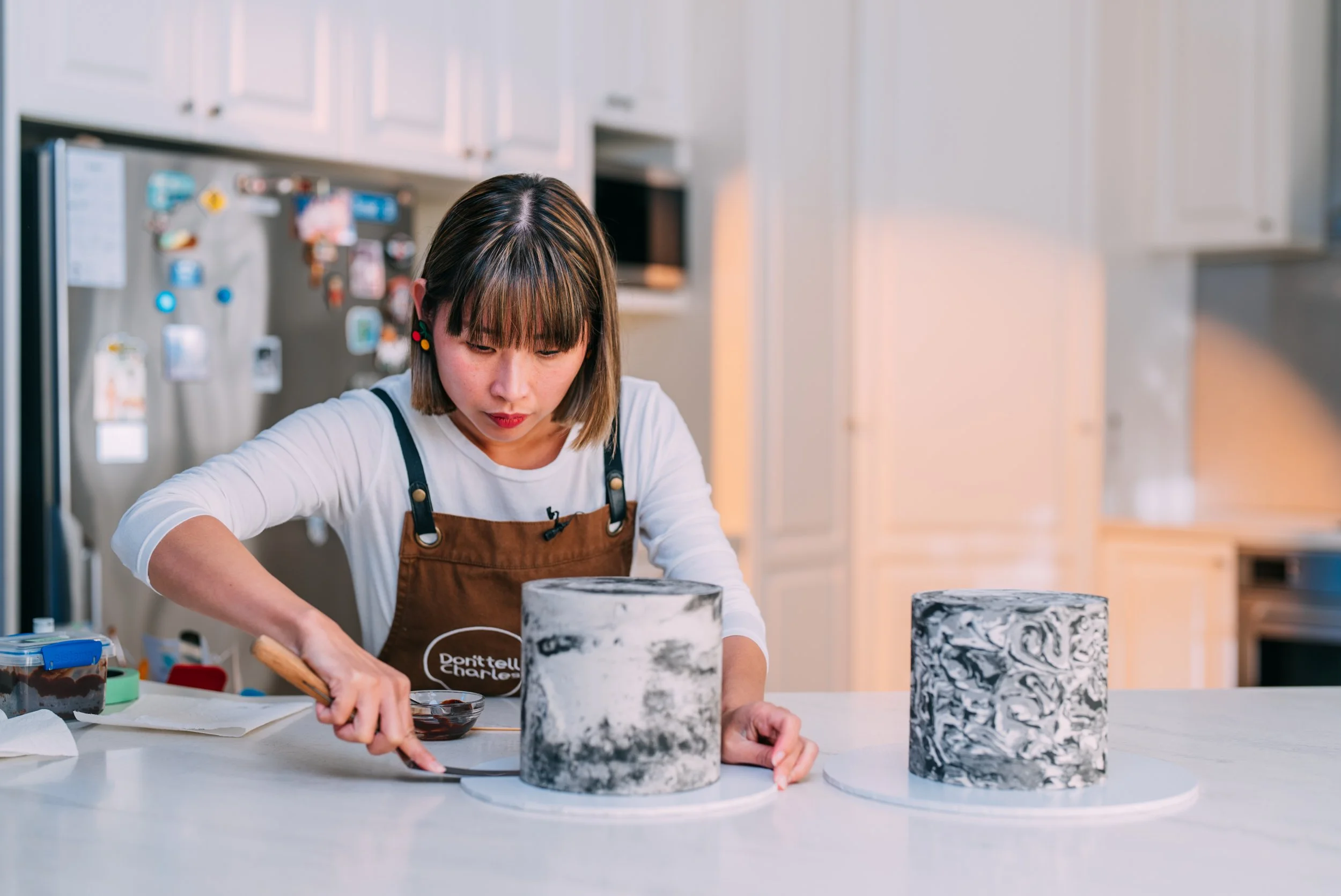 A woman decorated two cylindrical cakes with marbled black and white patterns on a white countertop in a modern kitchen.