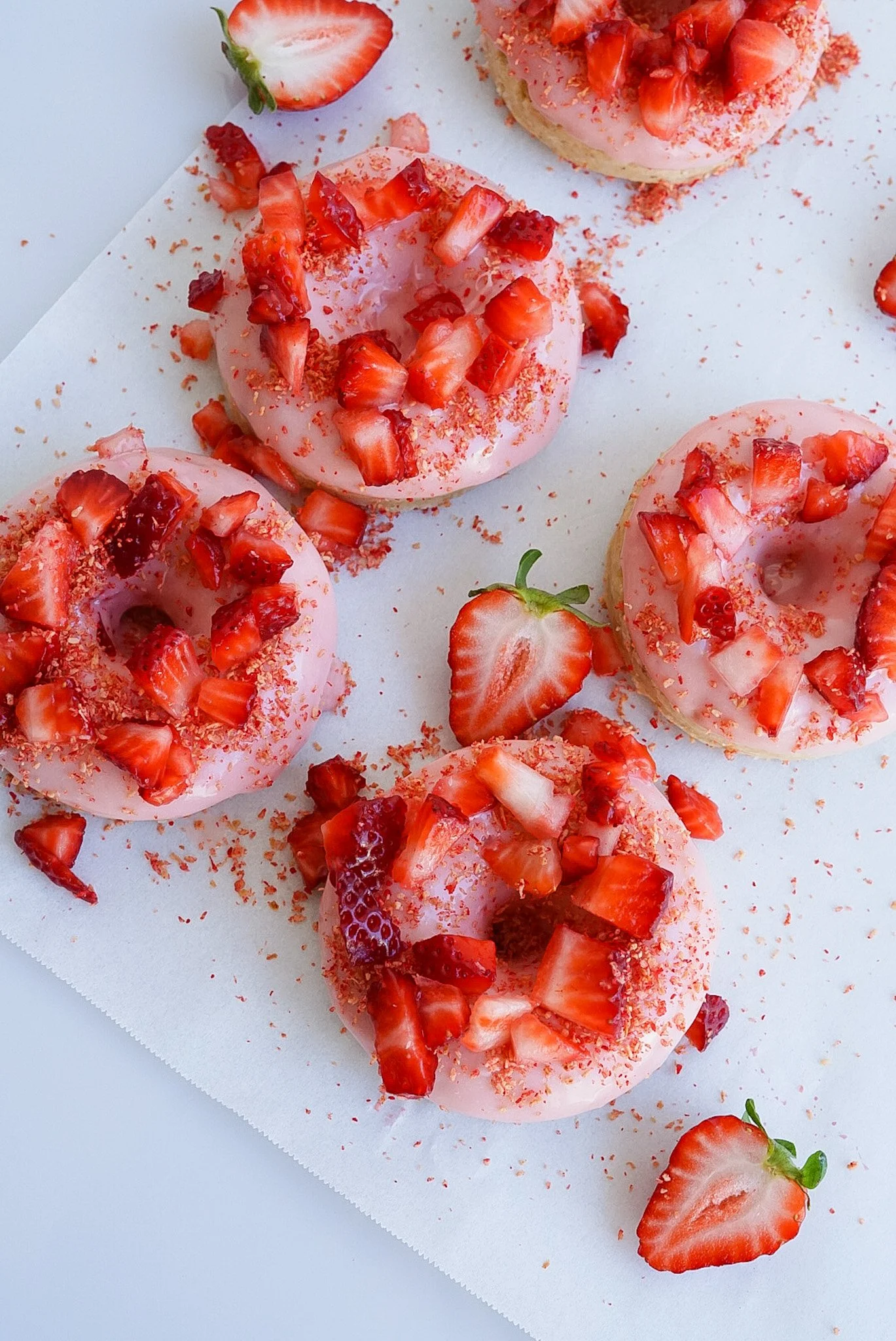 Pink frosted donuts topped with chopped strawberries and strawberry crumbs, with fresh strawberries beside them on white parchment paper.