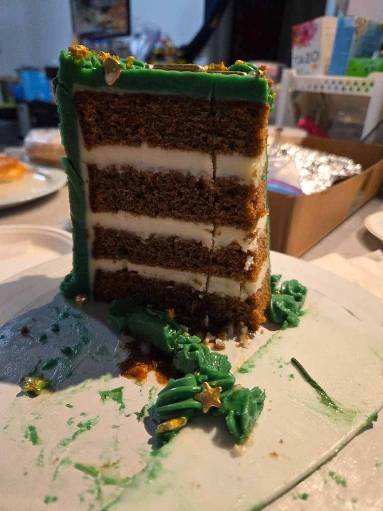 A partially eaten chocolate layer cake with white cream filling, decorated with green frosting and gold star ornaments, on a white cake board.