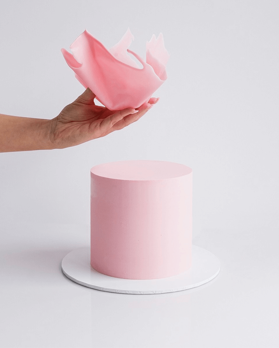 A hand holding a pink frosting flower cake decoration above a pink frosted cake on a white circular cake board against a white background.