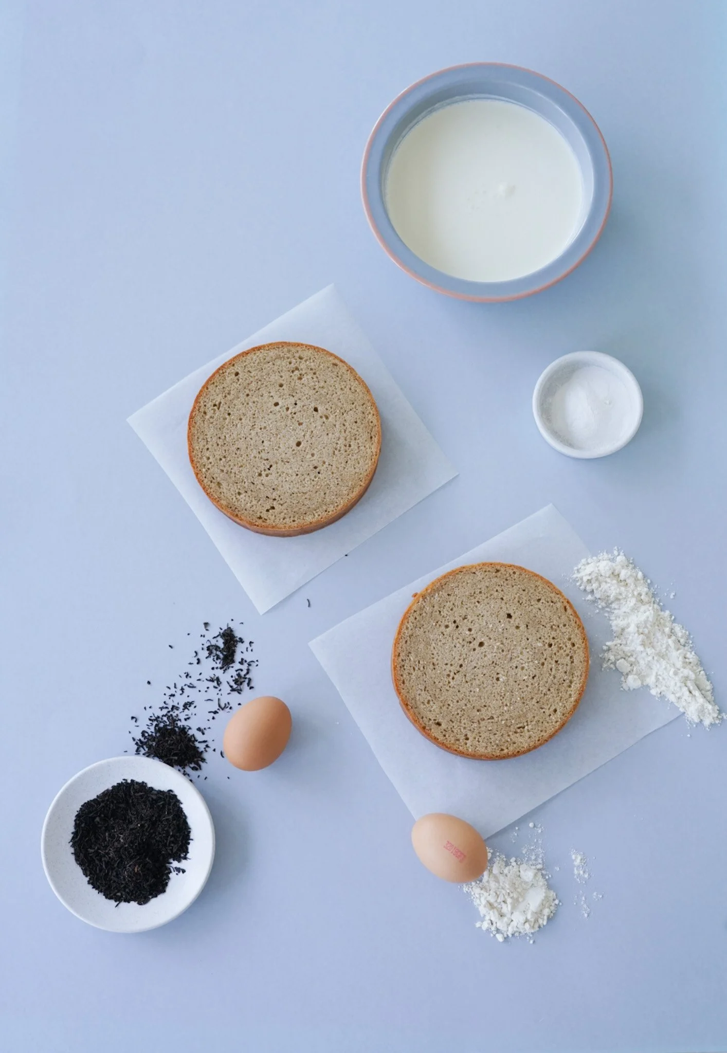 Ingredients for baking, including two vanilla sponge cakes on parchment paper, eggs, black tea leaves, flour, milk, salt, and baking powder on a white surface.