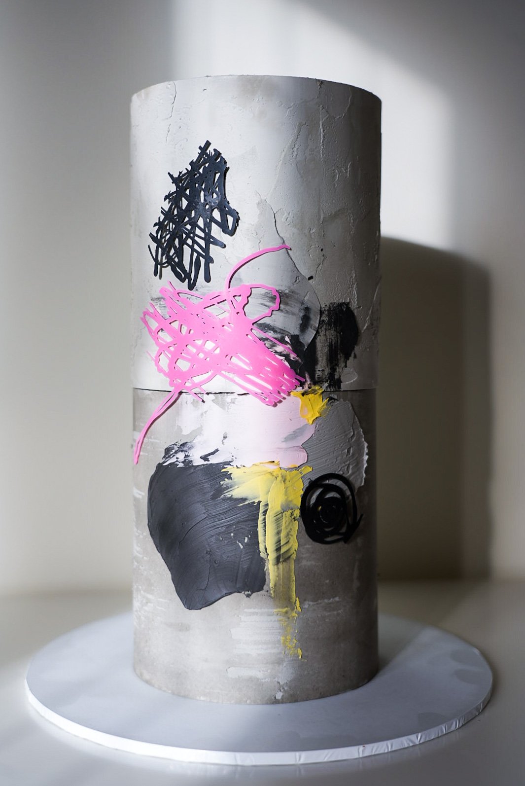 A cylindrical sculpture with a textured gray surface, decorated with abstract black, pink, yellow, and white paint strokes and line drawings, standing on a white circular base.