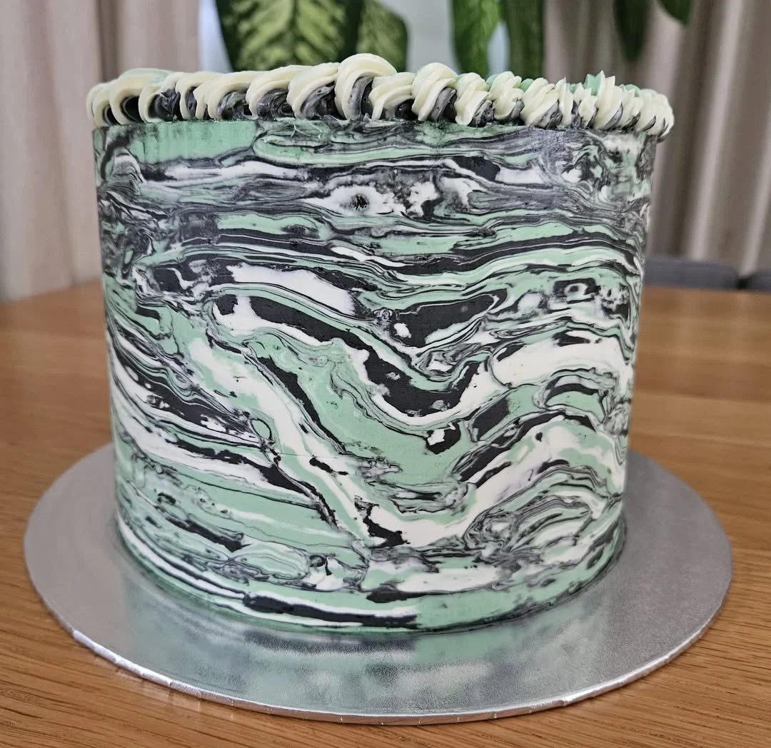 A cake with black, white, and green marbled swirl icing, decorated with a piped border along the top edge, placed on a silver cake board on a wooden table.