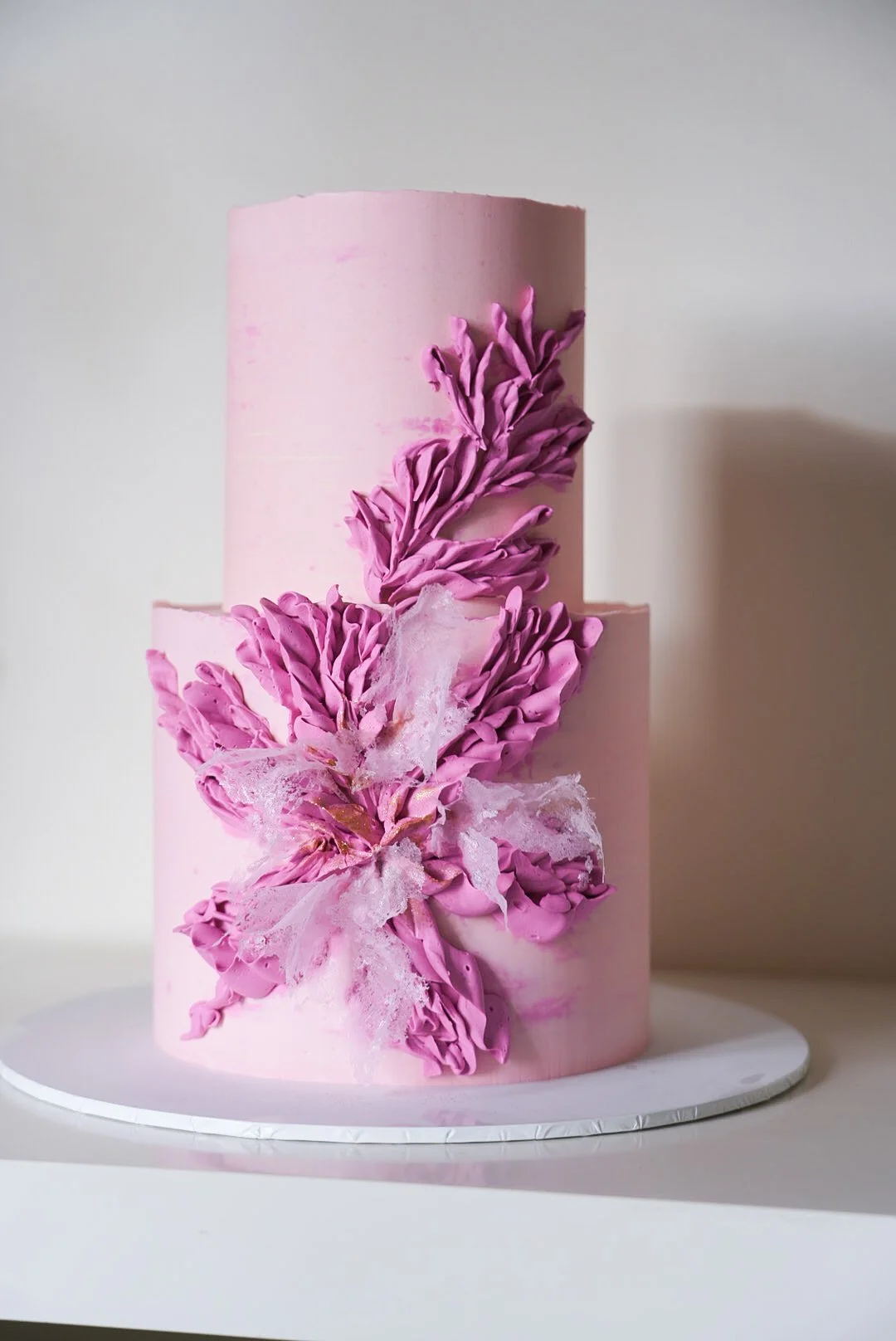 A two-tier pink cake decorated with ruffled pink flowers and delicate white sugar web accents.