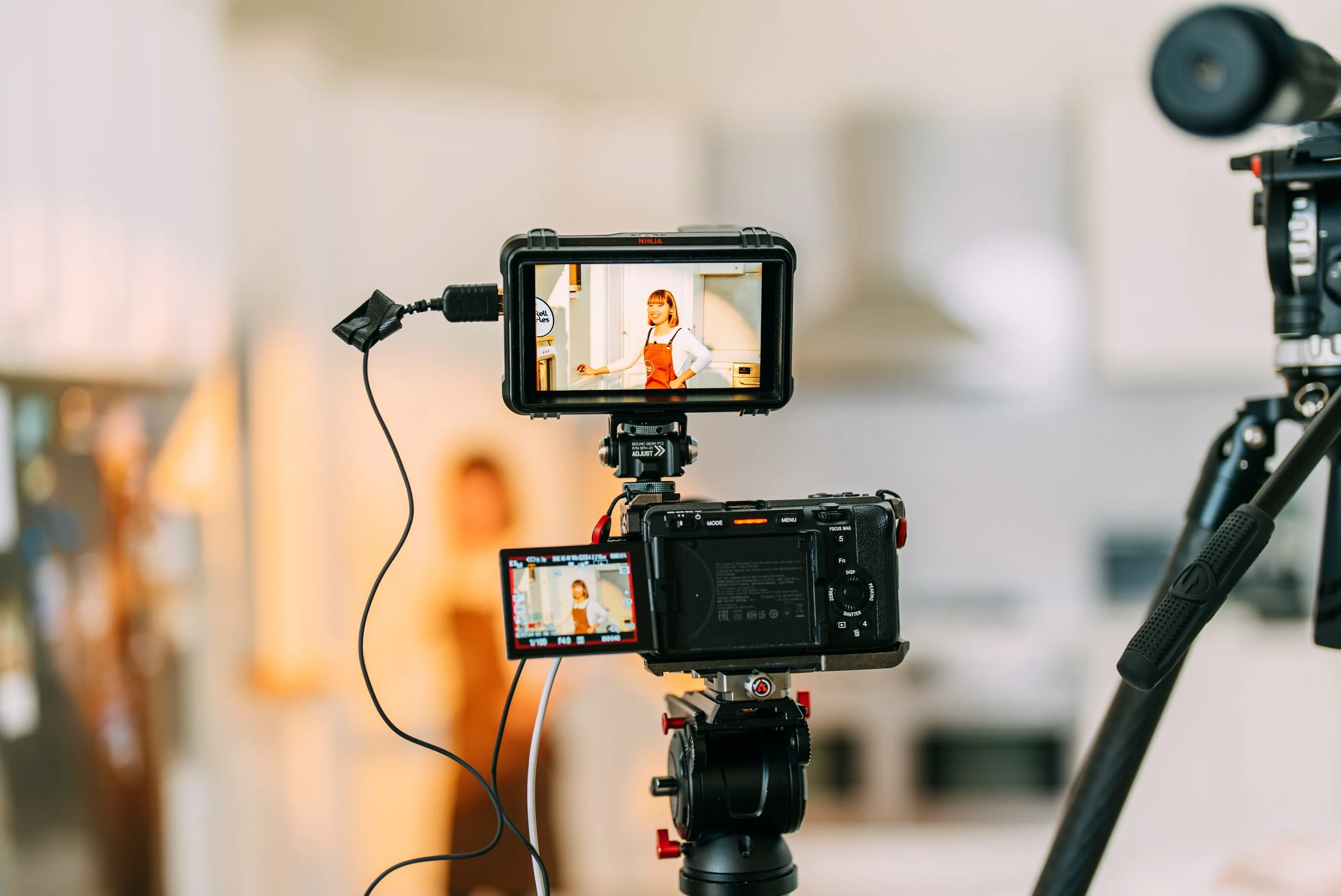 Camera setup capturing a woman in a kitchen. The camera's display shows a smiling woman wearing an orange apron over a white shirt, standing in a kitchen environment.