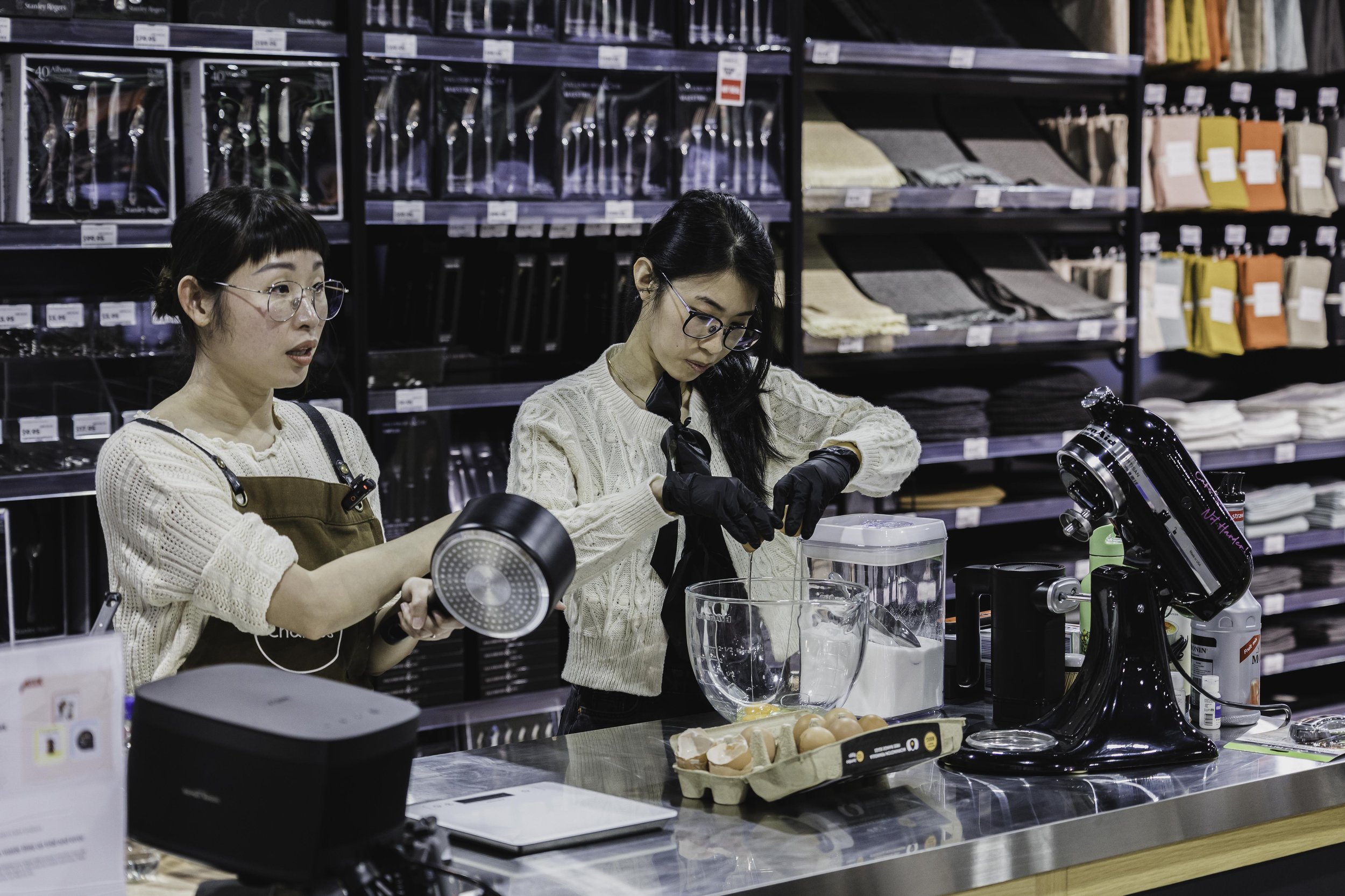 Two women preparing ingredients in a kitchen section of a store, surrounded by shelves of products.