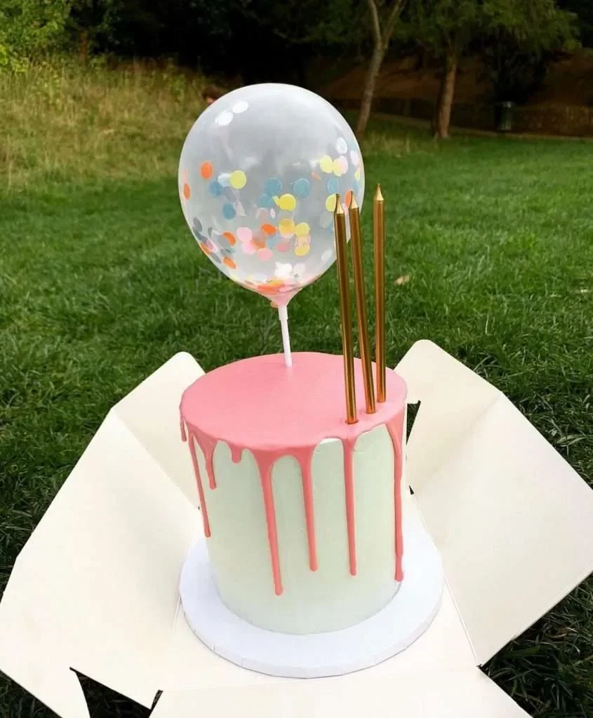 A birthday cake with pink dripping icing, a clear balloon with colorful confetti, and three gold candles outdoors on a grassy field.