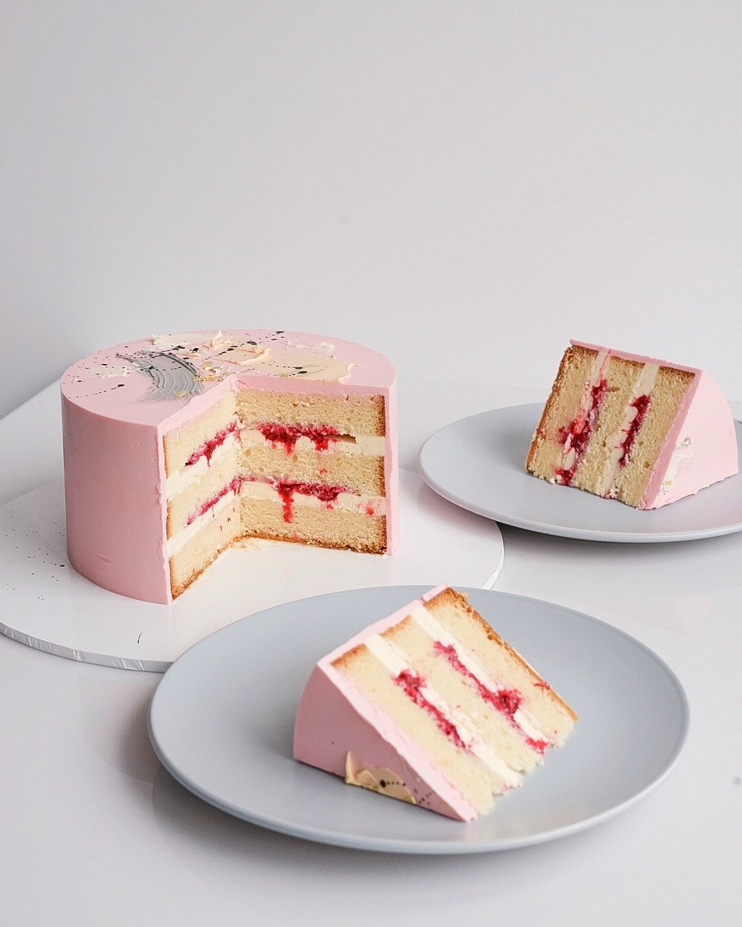 Pink layered cake with white and red filling, decorated with abstract painted designs, with a slice cut and placed on a plate, and another slice on a white cake stand.