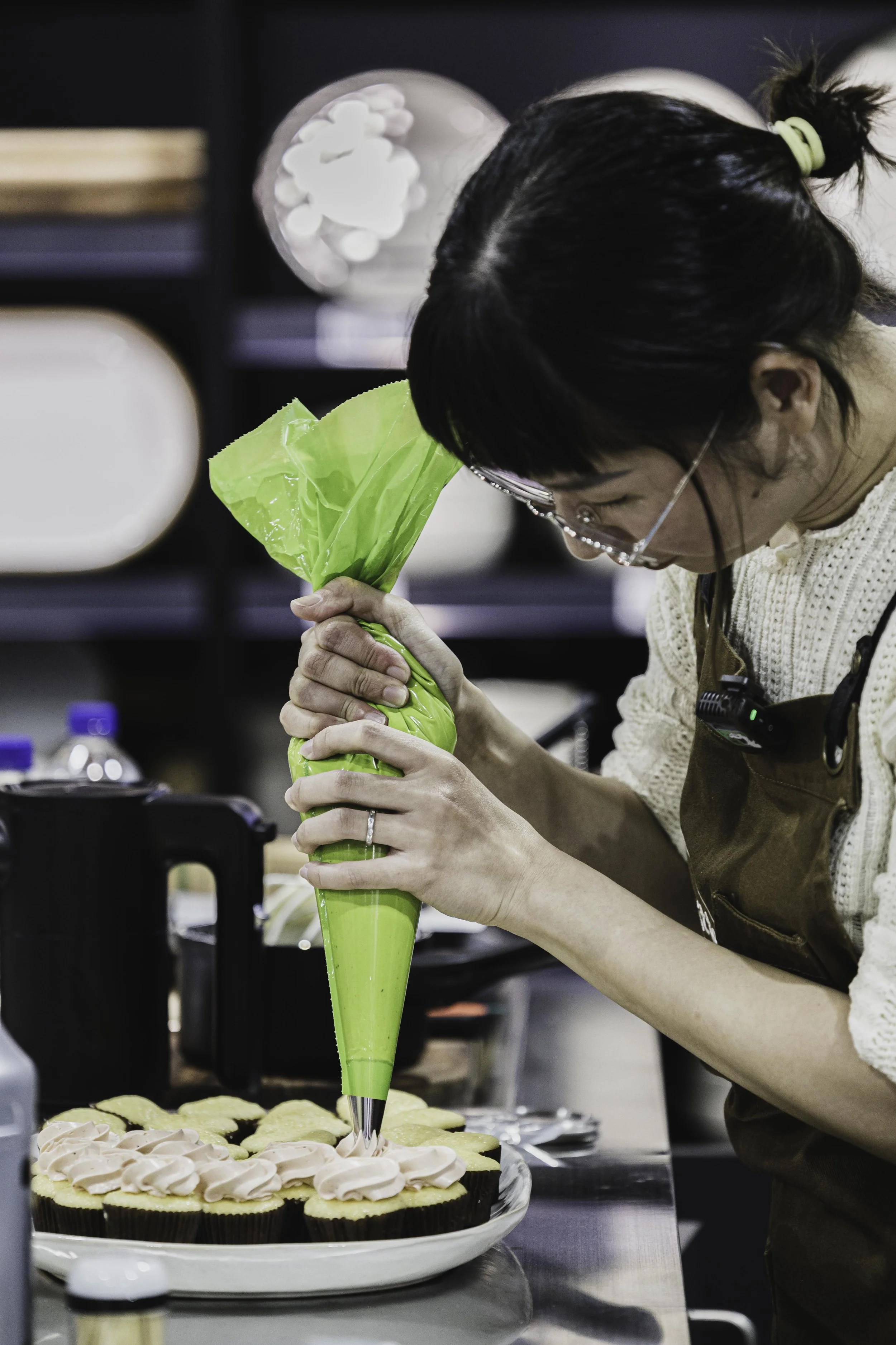 A woman in glasses and a brown apron decorates cupcakes with pink frosting using a large icing pastry bag in a kitchen or bakery setting.