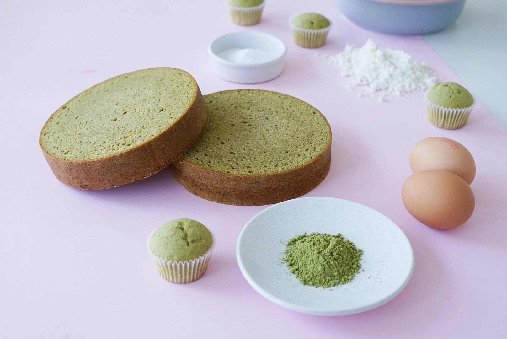 Matcha sponge cake ingredients on a pink surface, includes two sponge cake layers, matcha powder, eggs, matcha cupcakes, whipped cream, and flour.