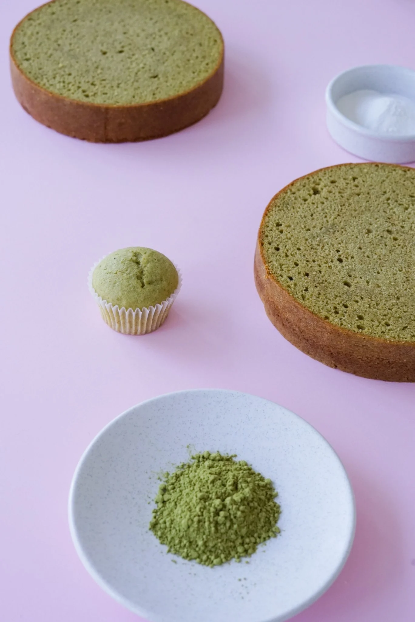 Matcha-flavored cake with a green matcha powder, a matcha cupcake, and a small bowl of whipped cream on a pink surface.