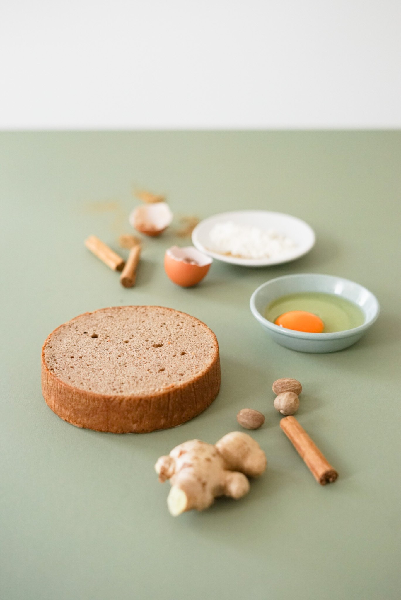 Raw ingredients on a green surface, including a slice of bread, ginger, cardamom pods, cinnamon sticks, an egg, and small dishes with flour and egg.