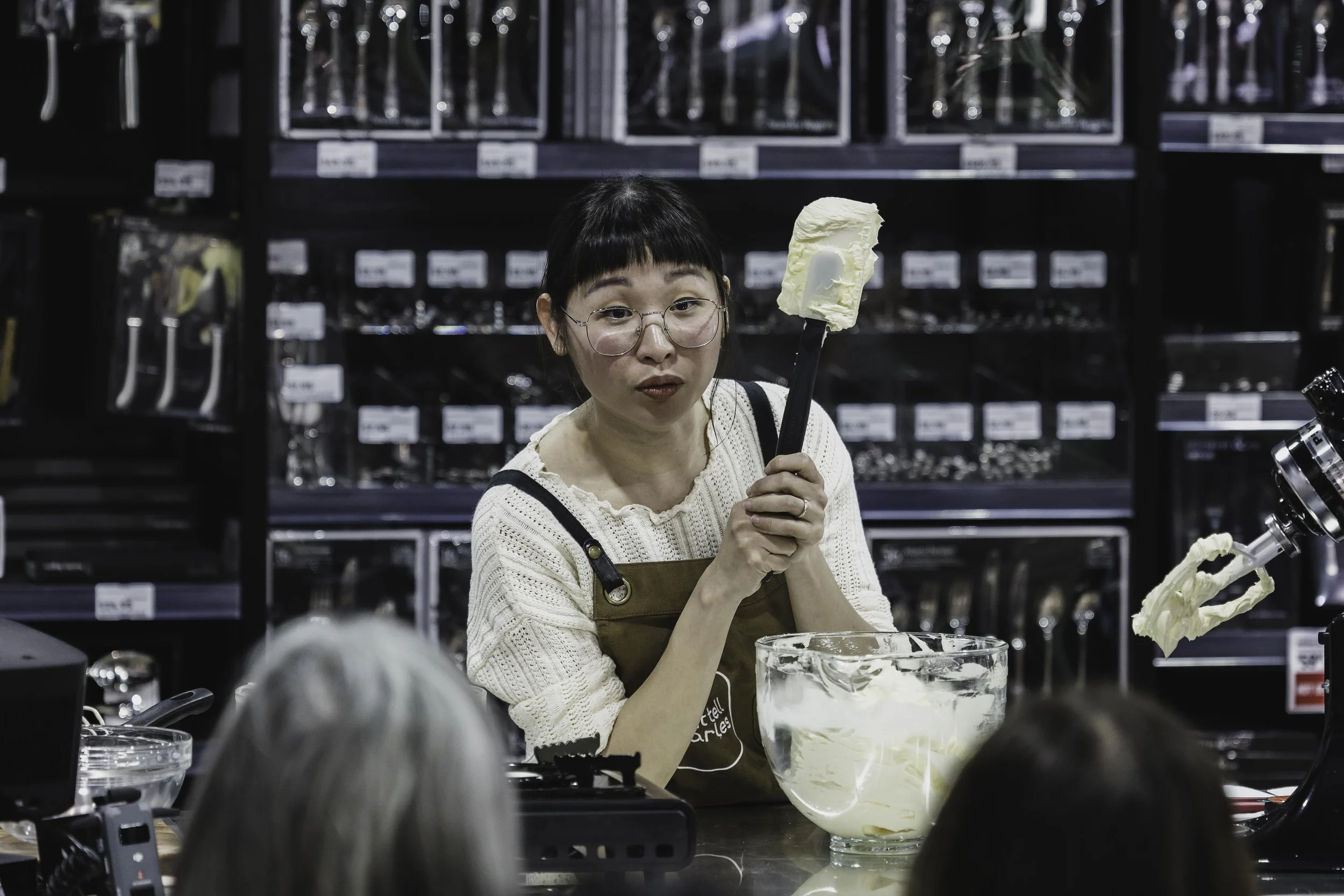 A woman with glasses and dark hair in a bob, wearing a beige sweater and brown apron, holding a spatula with butter in a kitchen or store setting, with baking supplies and shelves in the background.