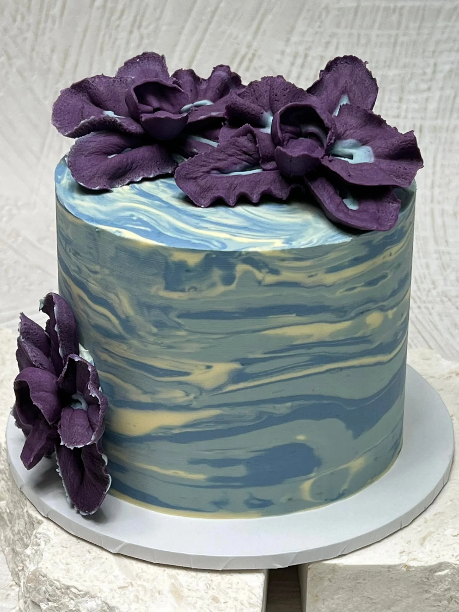 A marbled cake with purple flowers on top and to the side, placed on a white cake board.
