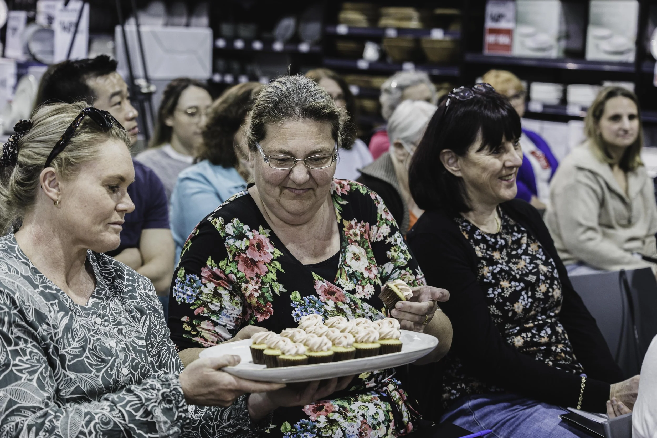 A group of women seated, one woman holding a tray of cupcakes, at an indoor event or gathering.