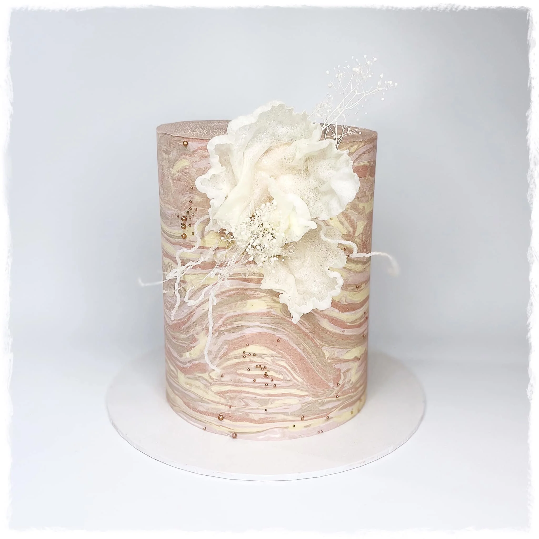A tall, cylindrical cake with a marbled pink, cream, and gold exterior, decorated with white edible flowers and delicate white sprigs on top.