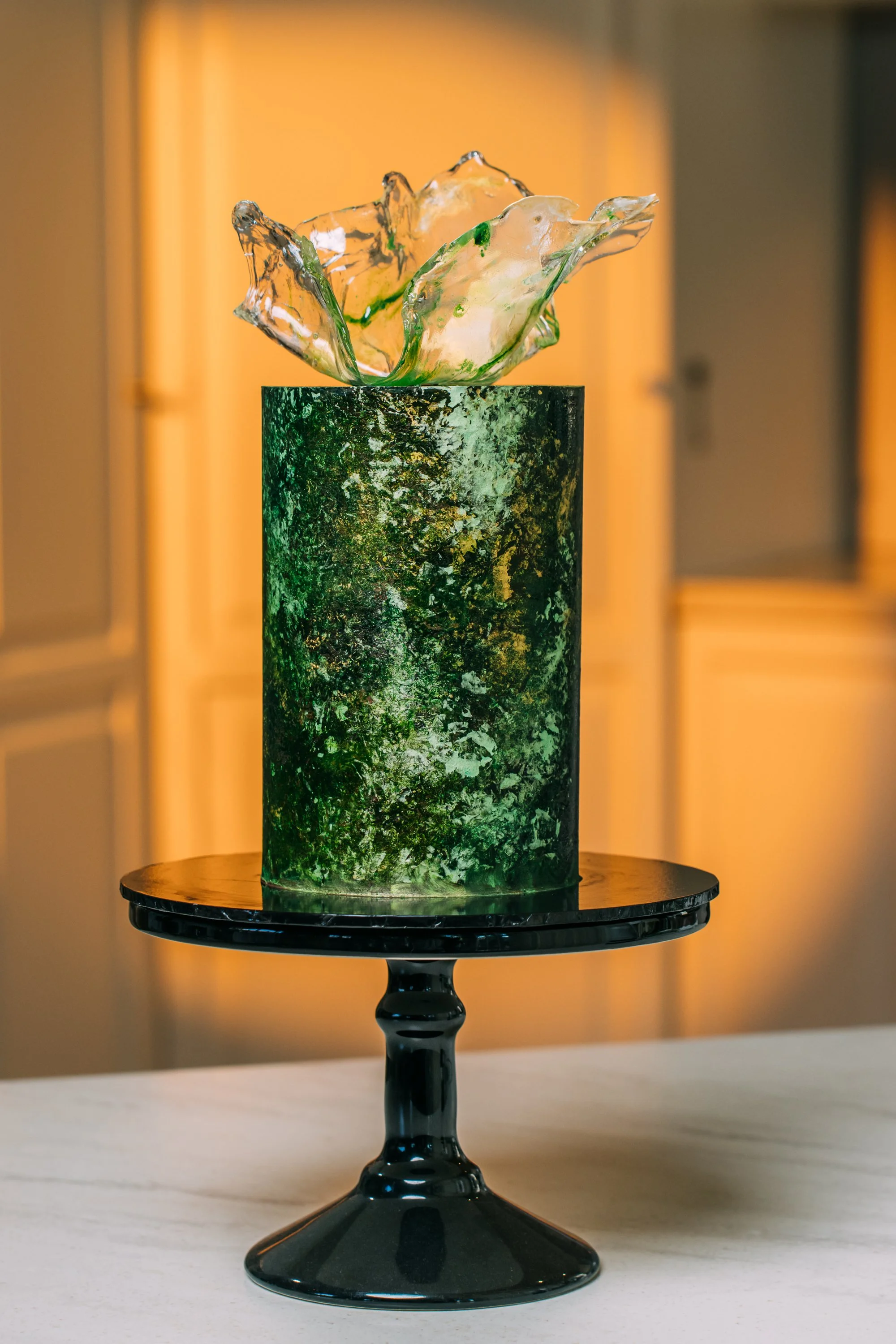 A decorative glass sculpture with abstract, flowing shapes on a green and black marbled pedestal, displayed on a black cake stand.
