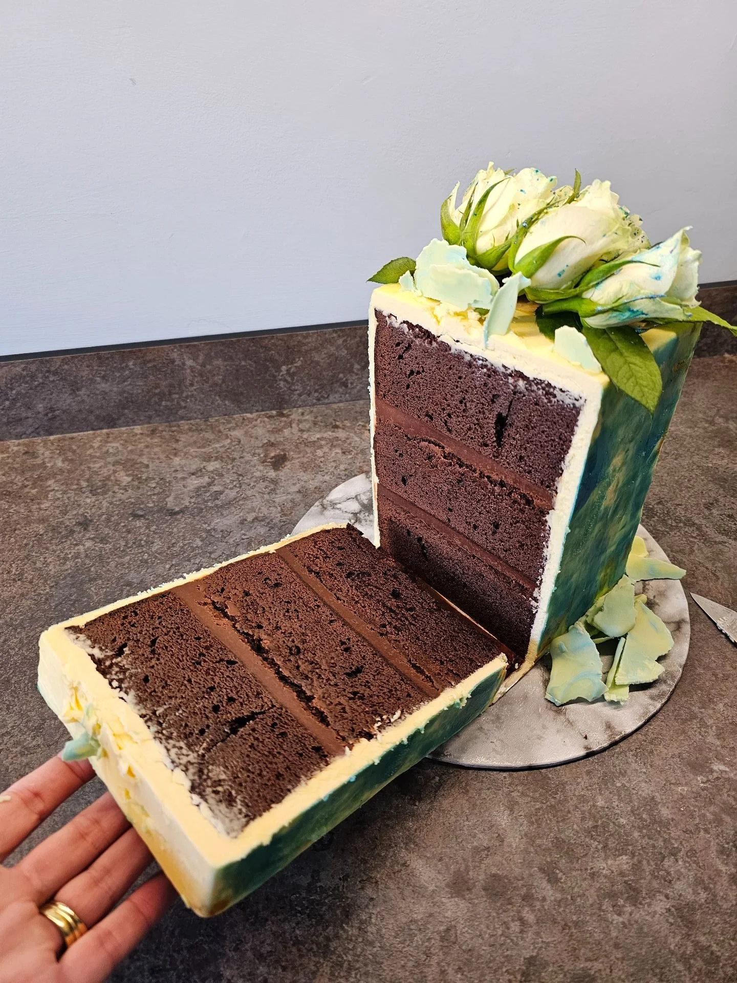 A rectangular layered chocolate cake with white frosting and green and yellow decorative flowers, with a slice being cut out and presented on a small round cake board.